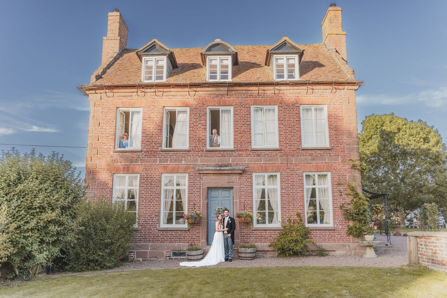 A couple stands in front of a grand historic red brick mansion with white trim and dormers under a blue sky.
