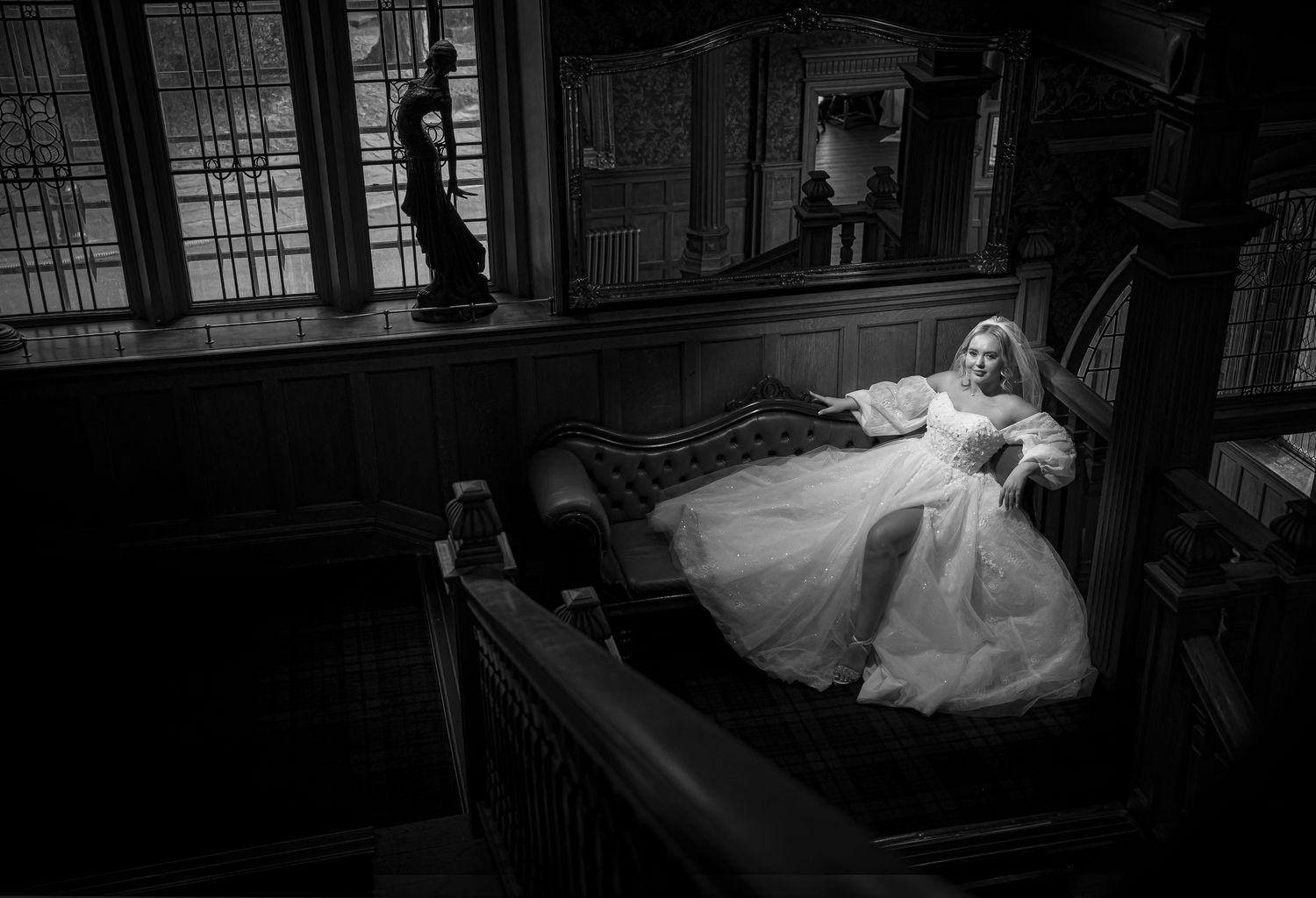 Bride in white gown reclining on a chaise lounge in a dark, moody room in black and white.