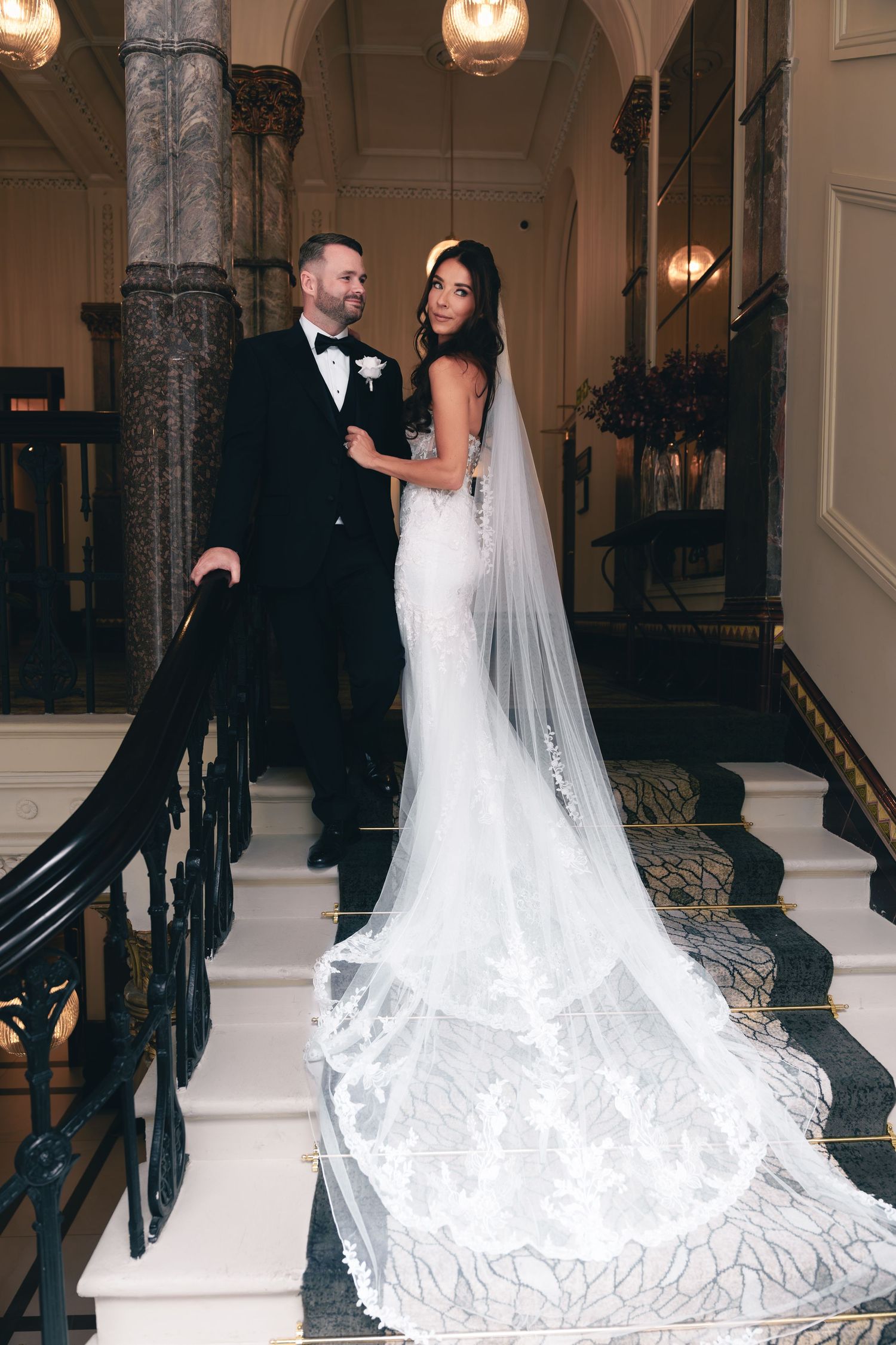 A wedding couple poses together in a grand marble hallway with elegant lighting and ornate architecture.