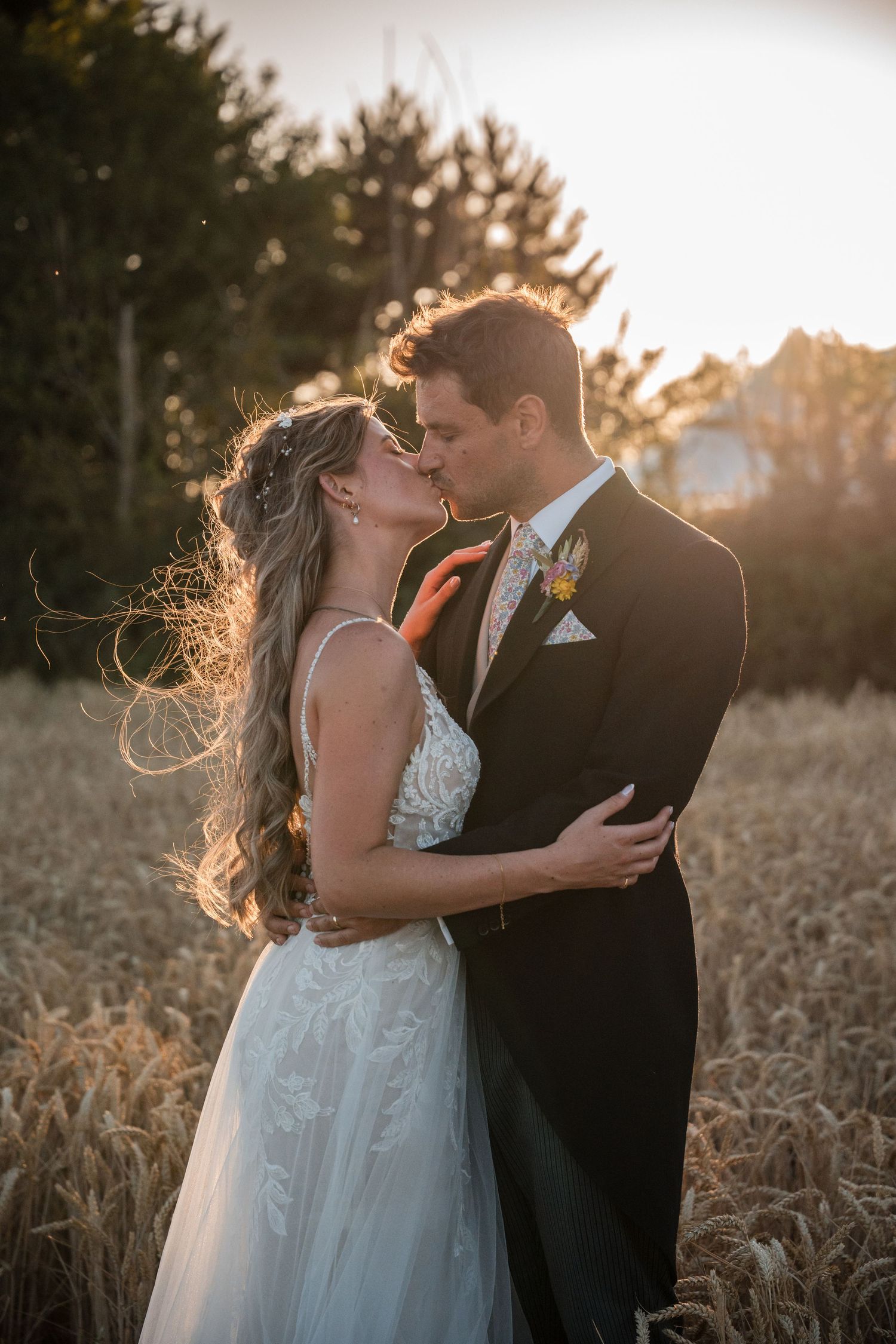 A romantic wedding photo captured at golden hour with backlit silhouettes embracing in a wheat field at sunset.