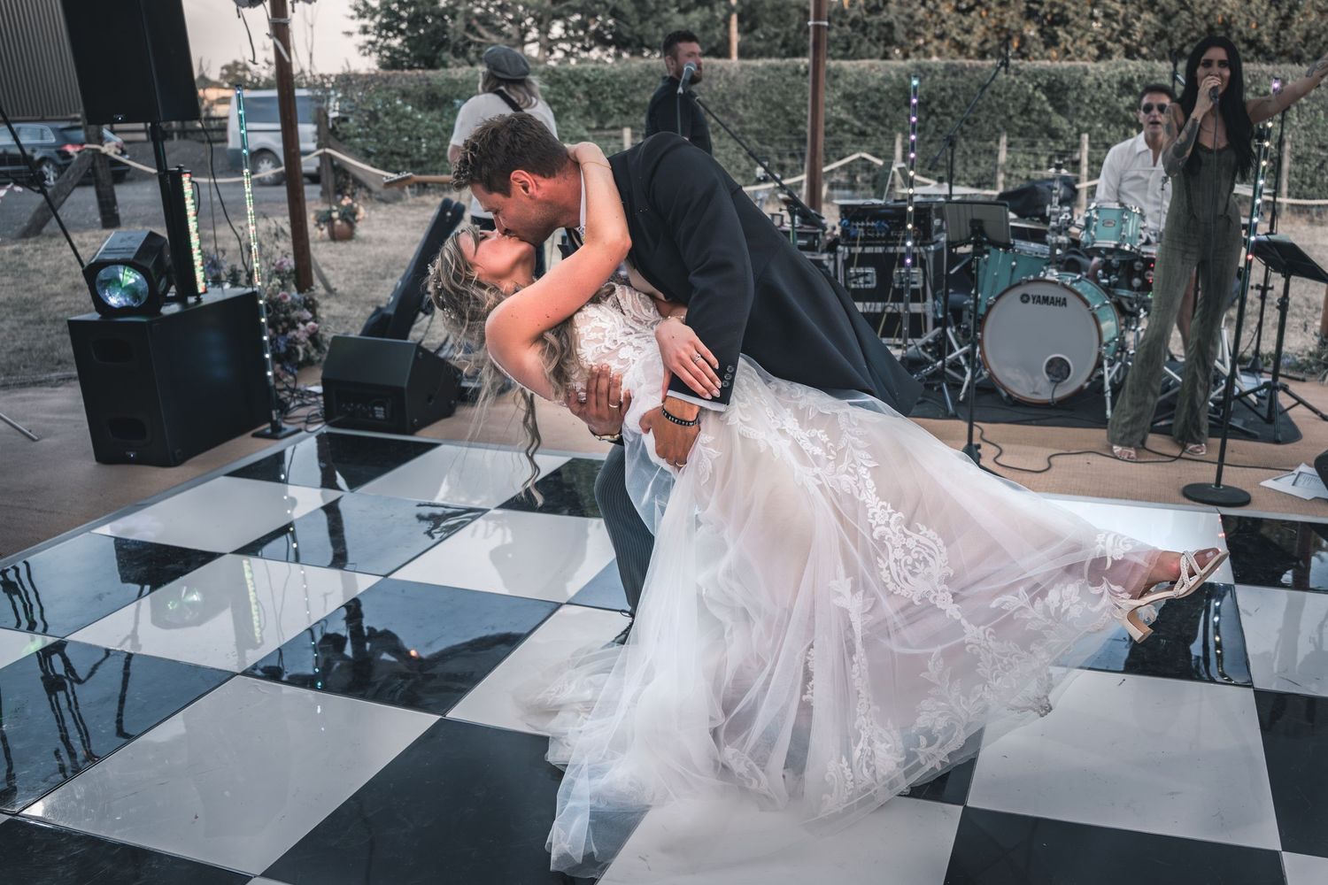 A romantic dip on a black and white checkered dance floor with a bride in lace gown and stage equipment in background.