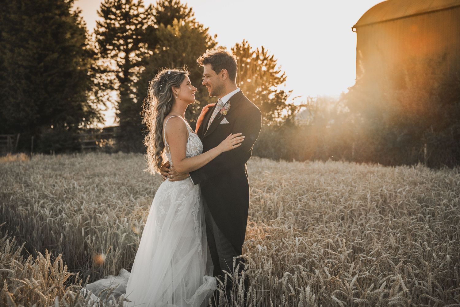 A couple shares an intimate embrace in a golden wheat field at sunset as warm light filters through trees.