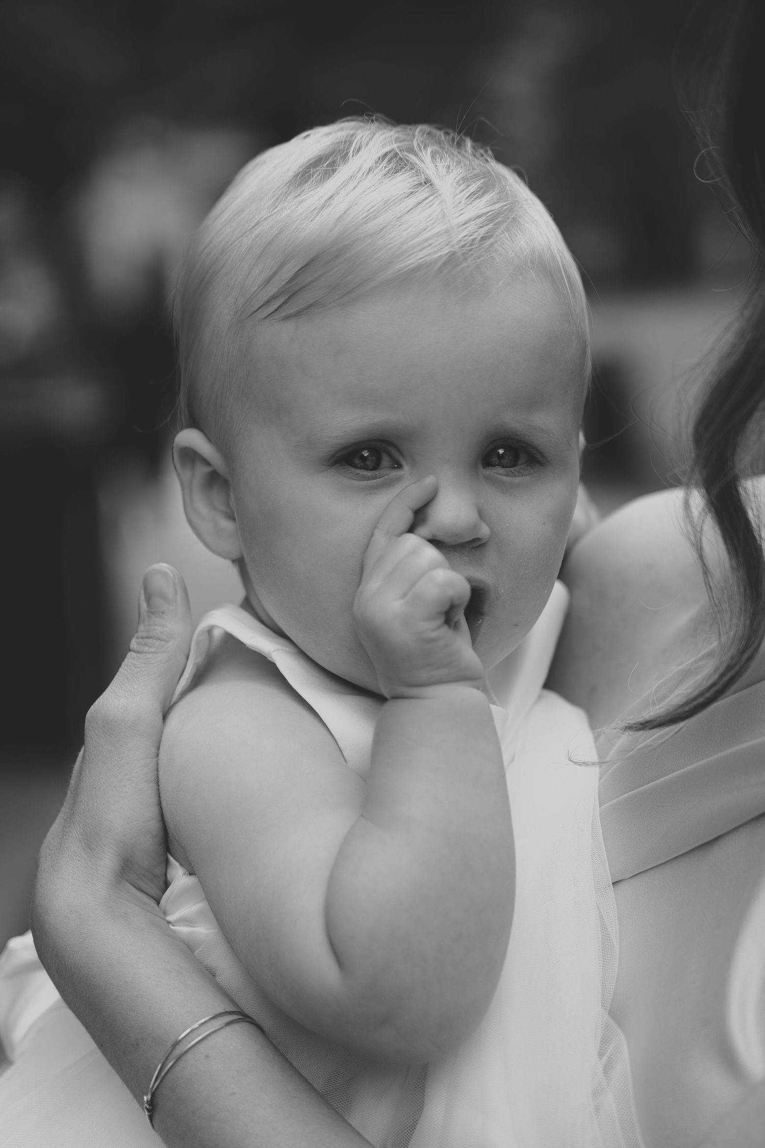A black and white portrait of a baby in white dress sucking their thumb while being held.