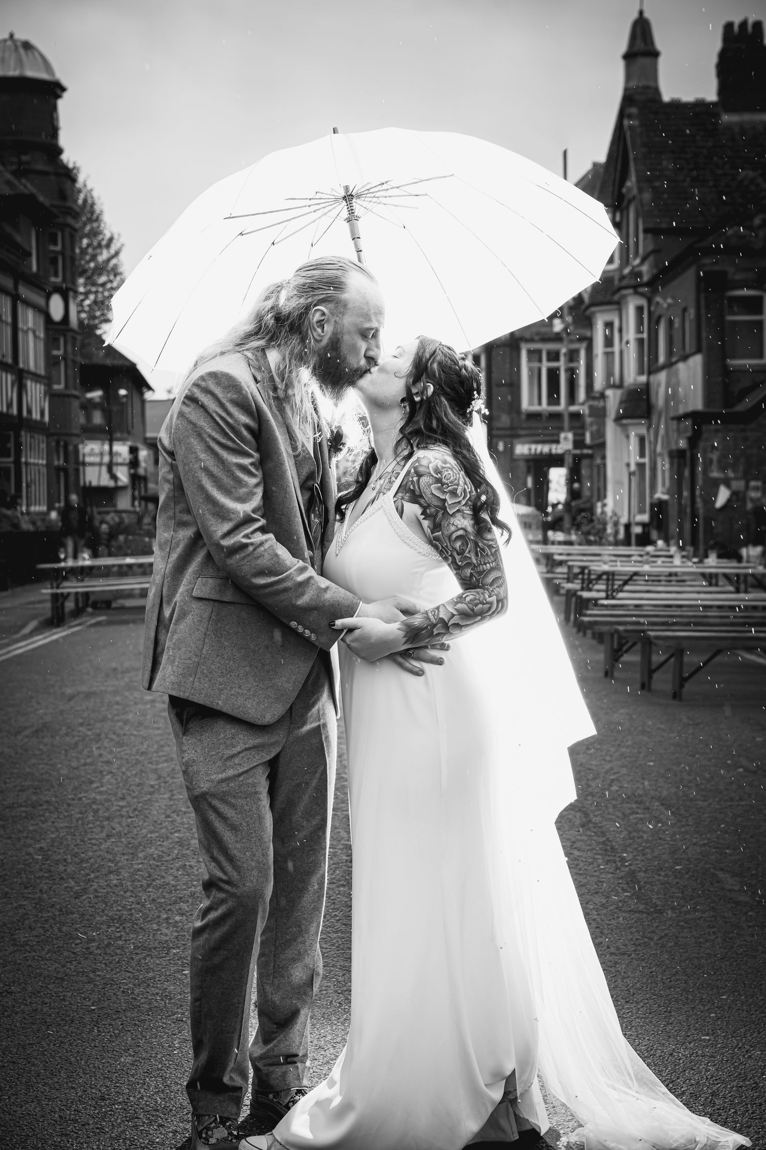 Black and white photo of a couple kissing under a white umbrella on a rainy street, bride in gown, groom in suit.