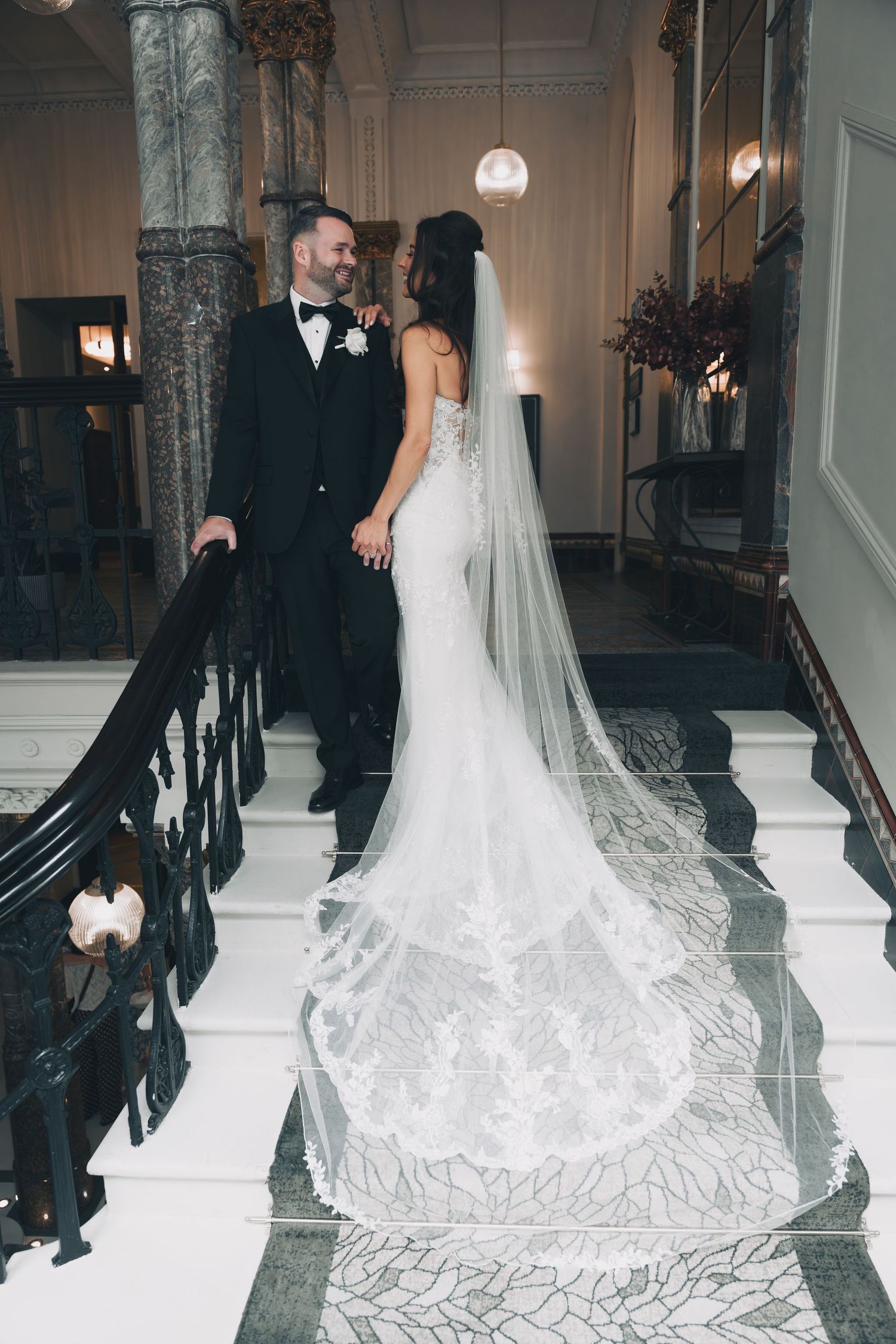 Bride in elegant lace gown with flowing cathedral veil walking down grand staircase at indoor wedding venue.