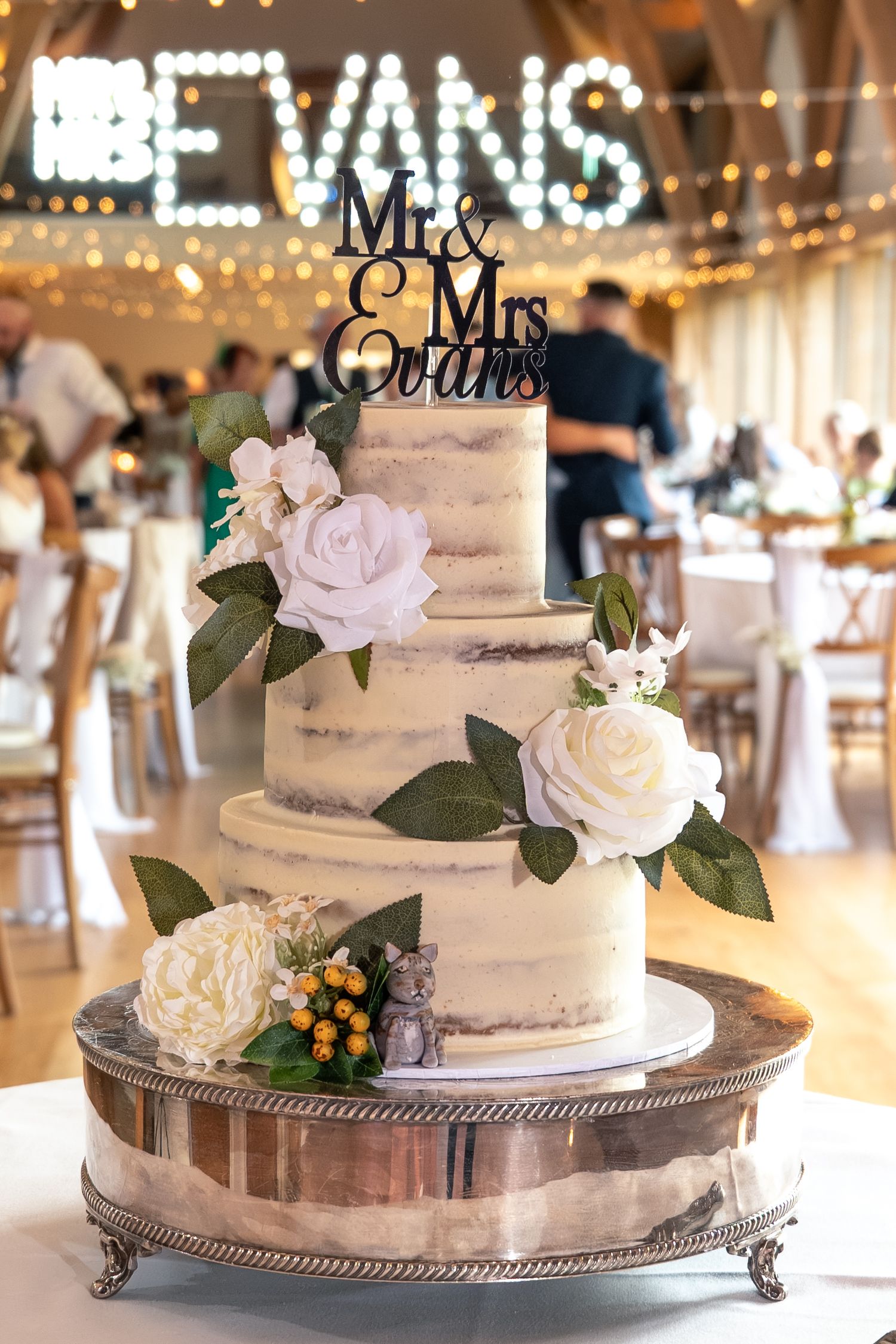 A three-tier white wedding cake decorated with roses sits beneath illuminated 'EVANS' signage.