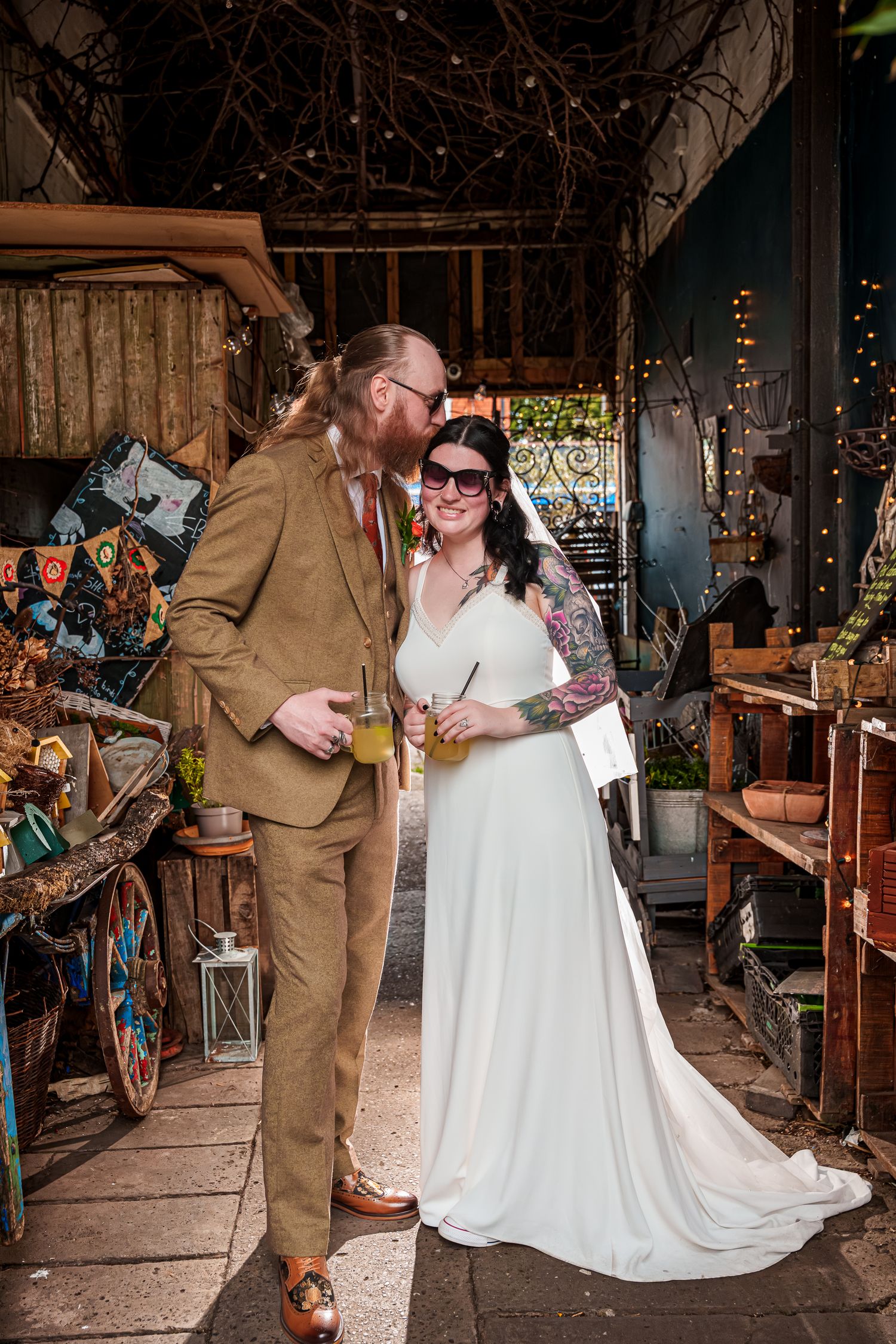 Bride and groom holding drinks share a kiss in a rustic, eclectic venue decorated with fairy lights and vintage decor.