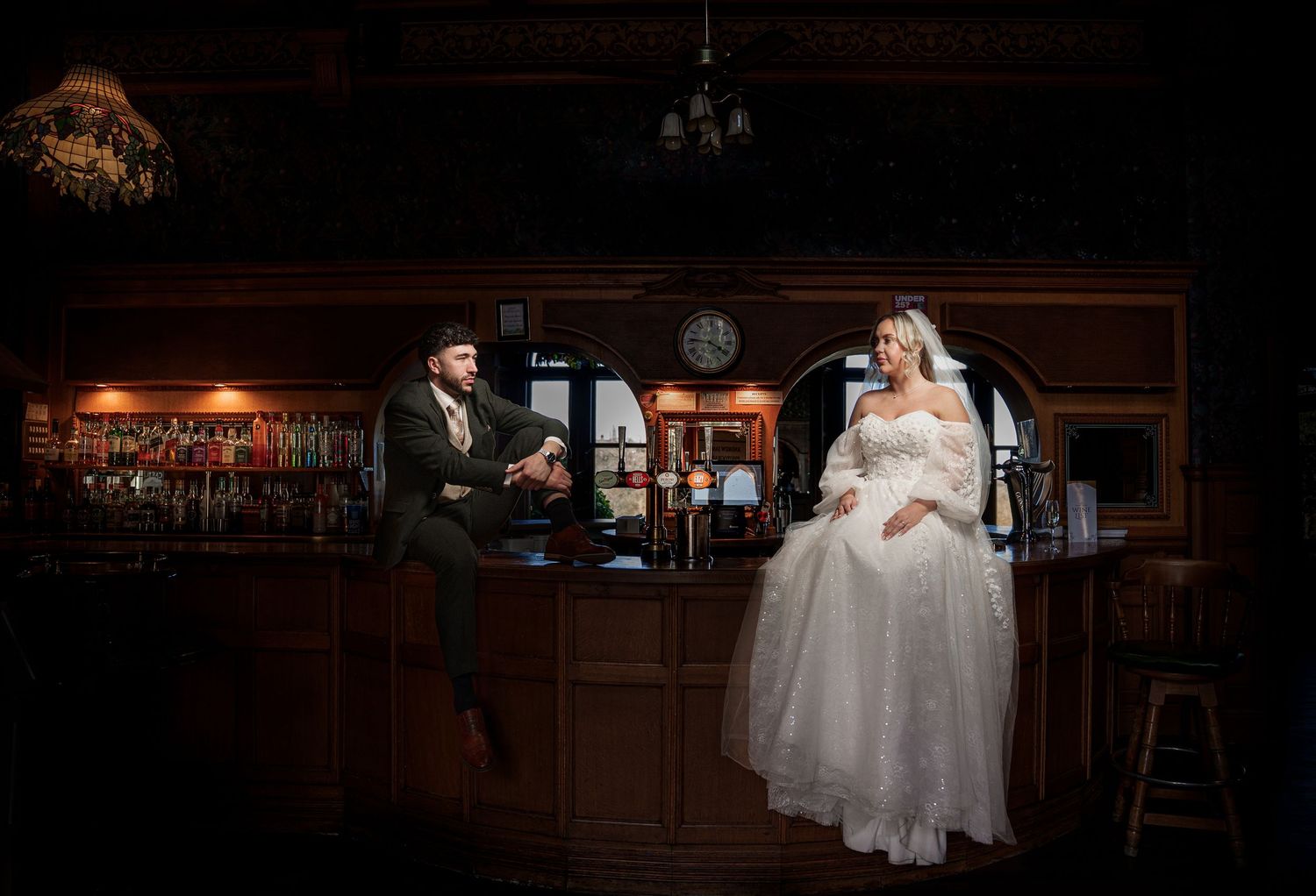 Bride and groom pose in elegant wooden bar setting, bride in white gown, groom in suit, warm romantic lighting.