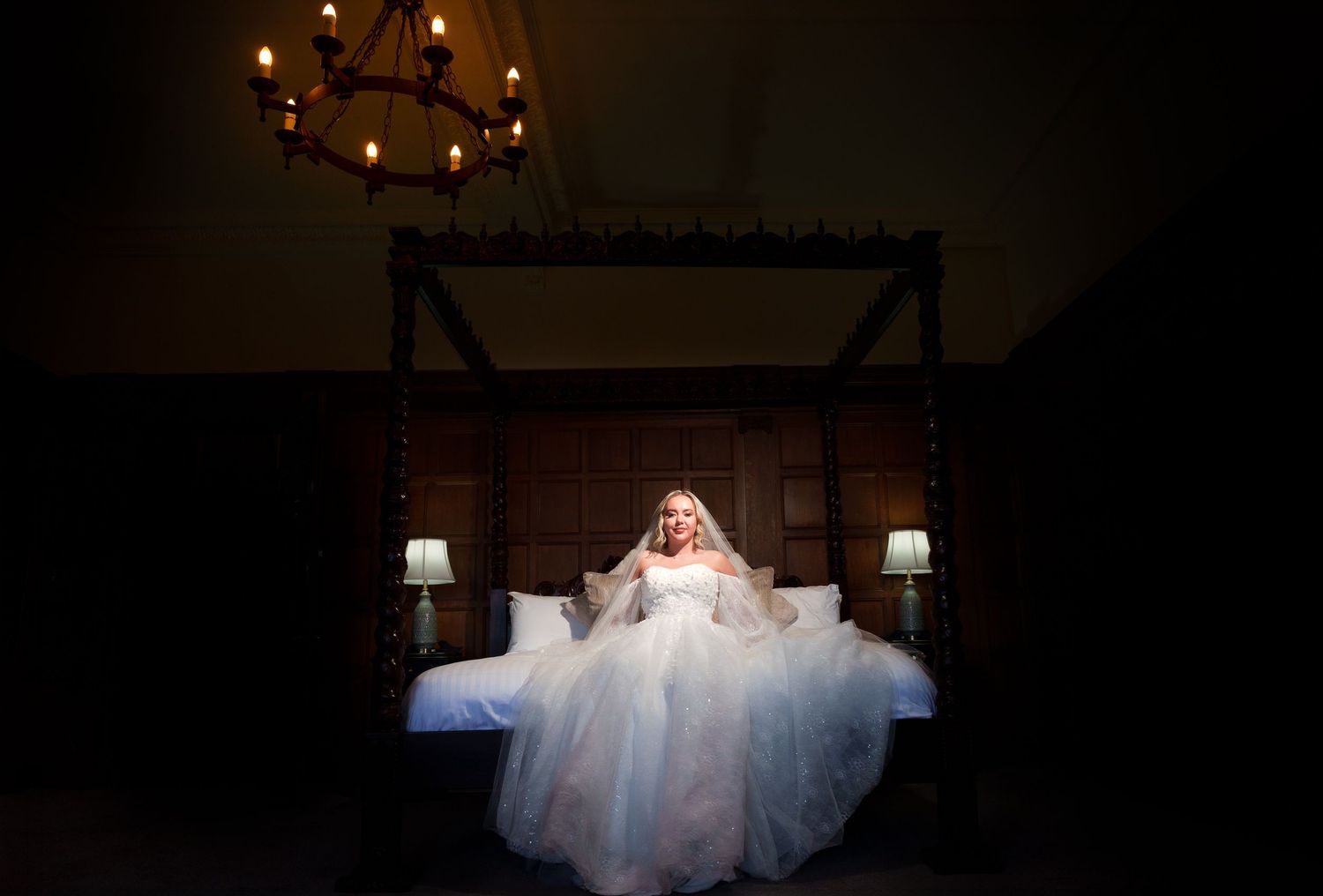 Bride in a flowing white wedding gown seated on a four-poster bed, illuminated by warm light in an elegant dark room.