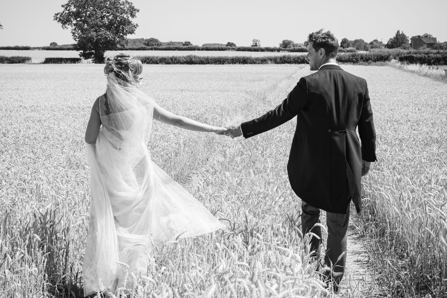 A couple walks hand in hand through a scenic wheat field captured in black and white.