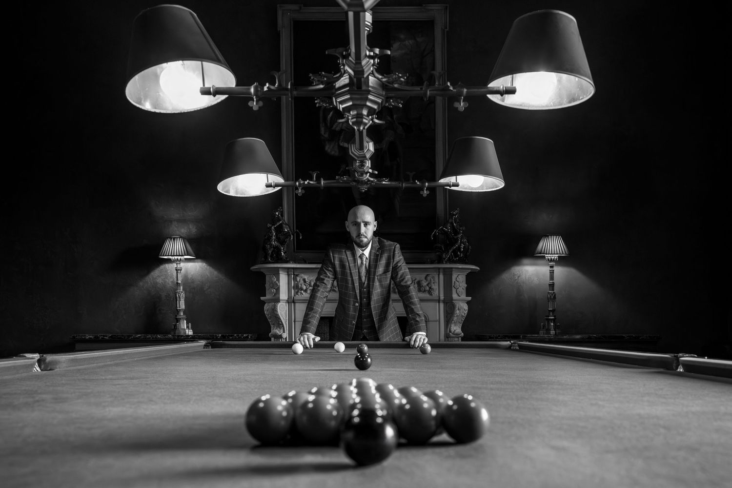 A dramatic black and white shot of pool balls set up on a billiards table under multiple hanging lamps.