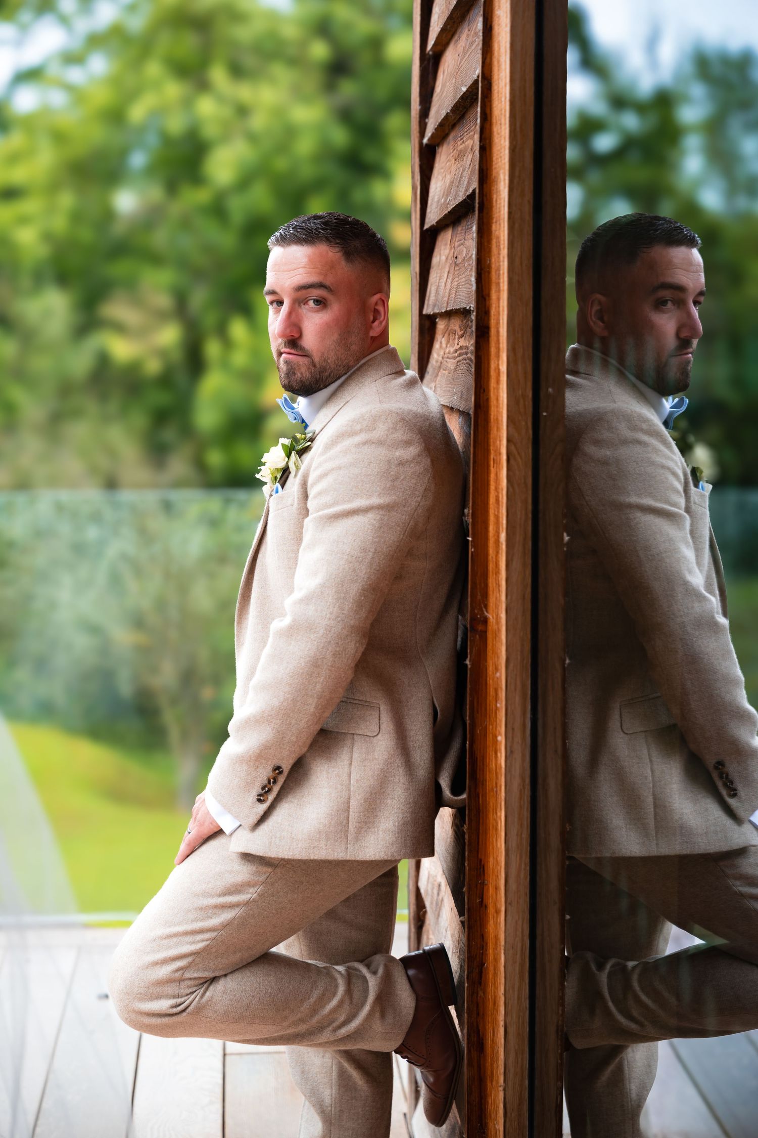 A groom in a tan suit poses against a wooden wall with his reflection visible.
The Mill Barns wedding venue