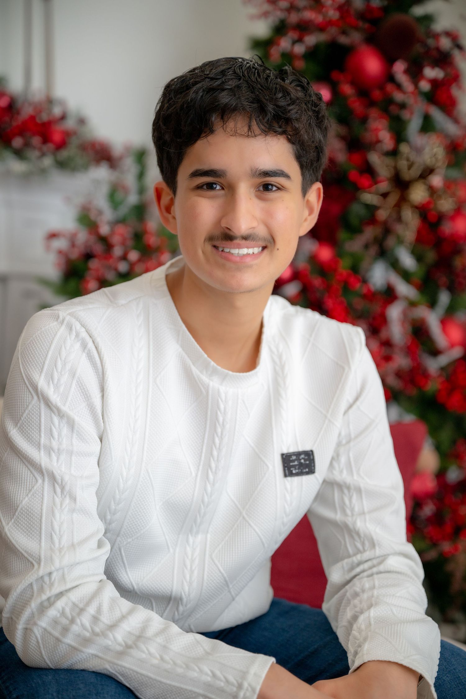 Smiling young man in white cable-knit sweater posing before red Christmas decorations.