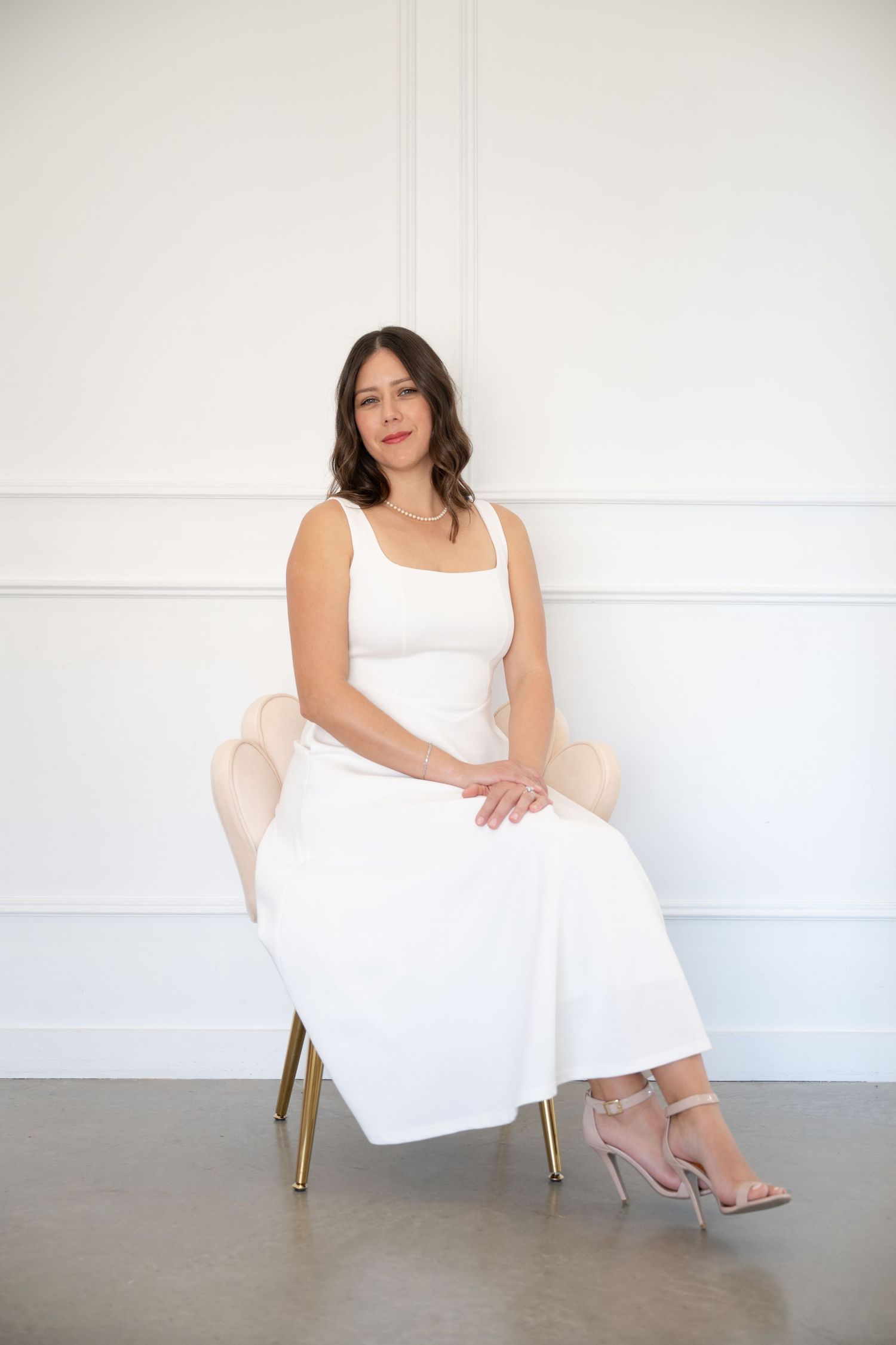 Woman in white sleeveless dress and heels sitting gracefully on a blush velvet chair against a white wall.