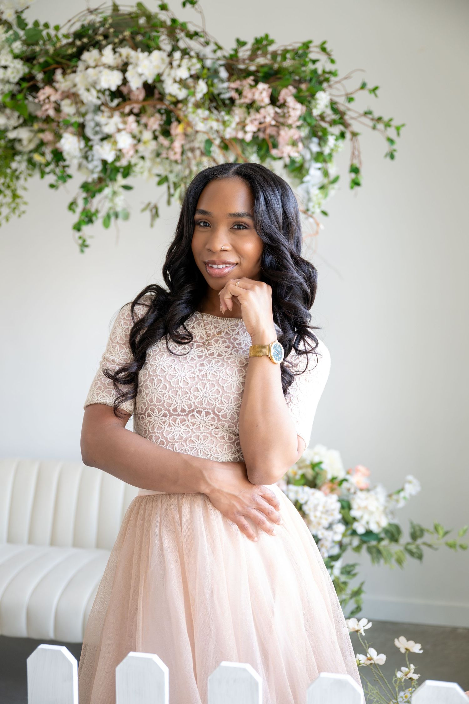 A woman in a white lace crop top and blush tulle skirt smiles beside a floral arrangement indoors.