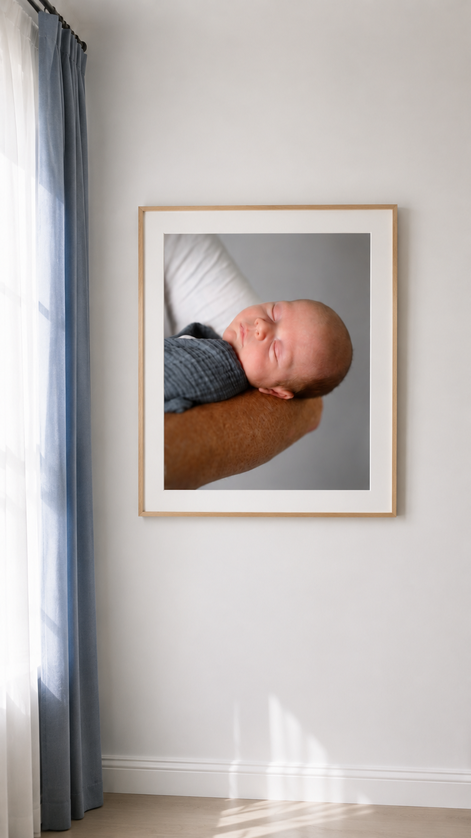 Framed newborn photo of a sleeping baby swaddled in blue, cradled in an adult's arm, displayed on a white wall.