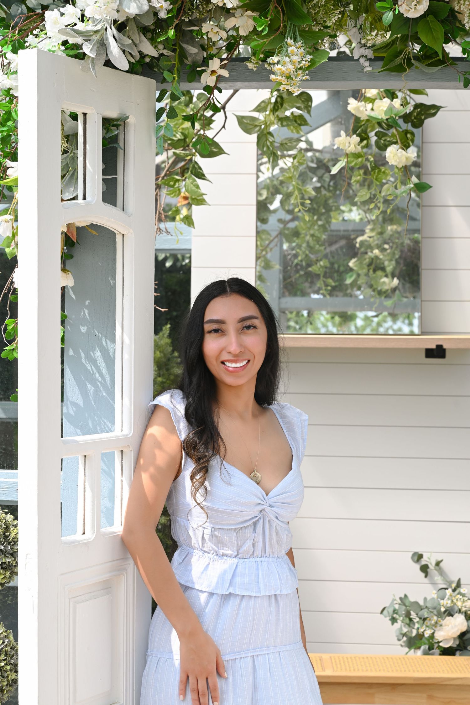 Young woman in light blue dress smiling beside a white door adorned with white flowers and greenery.