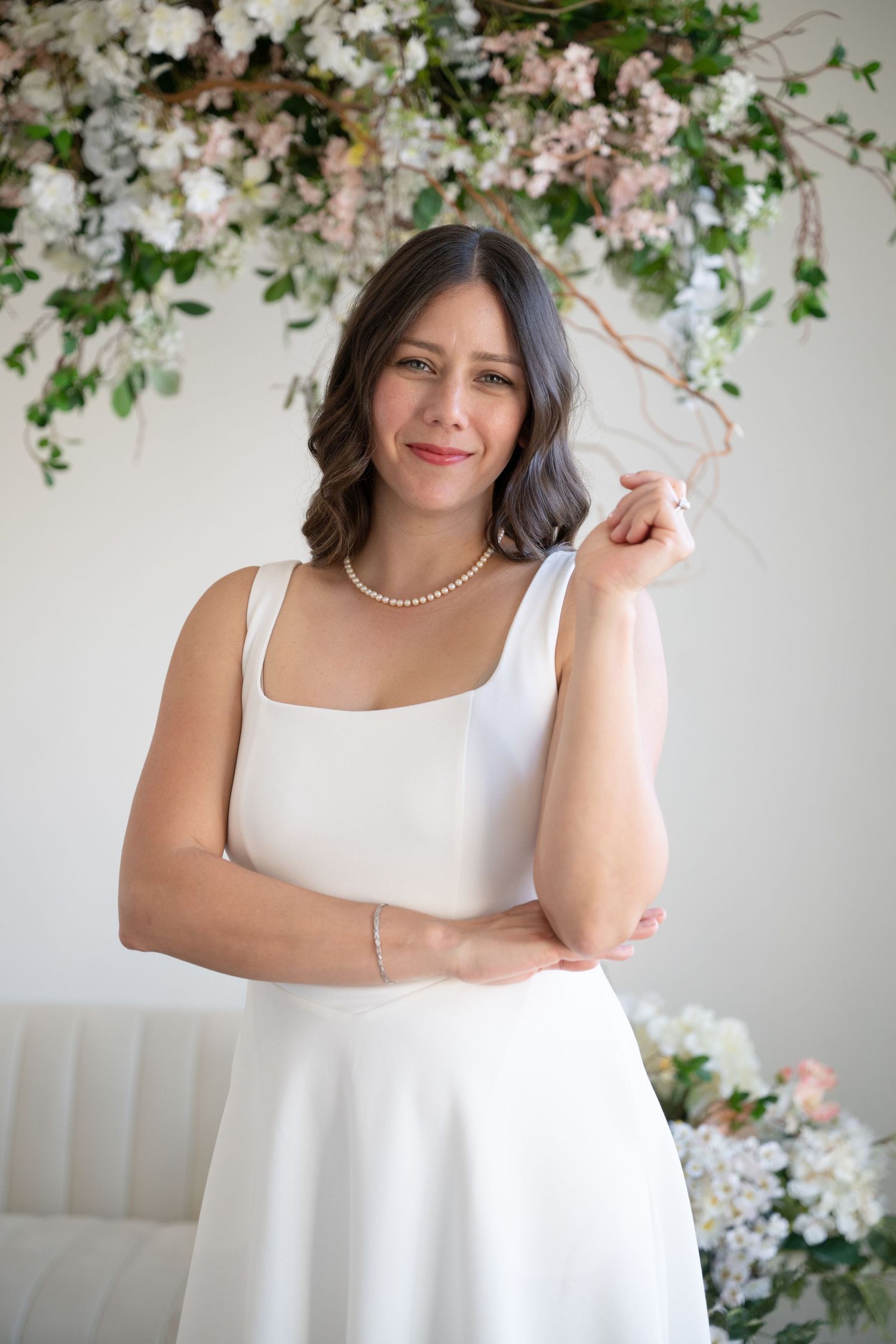 Woman in white dress with pearl necklace poses before floral arrangement of pink and white blooms.