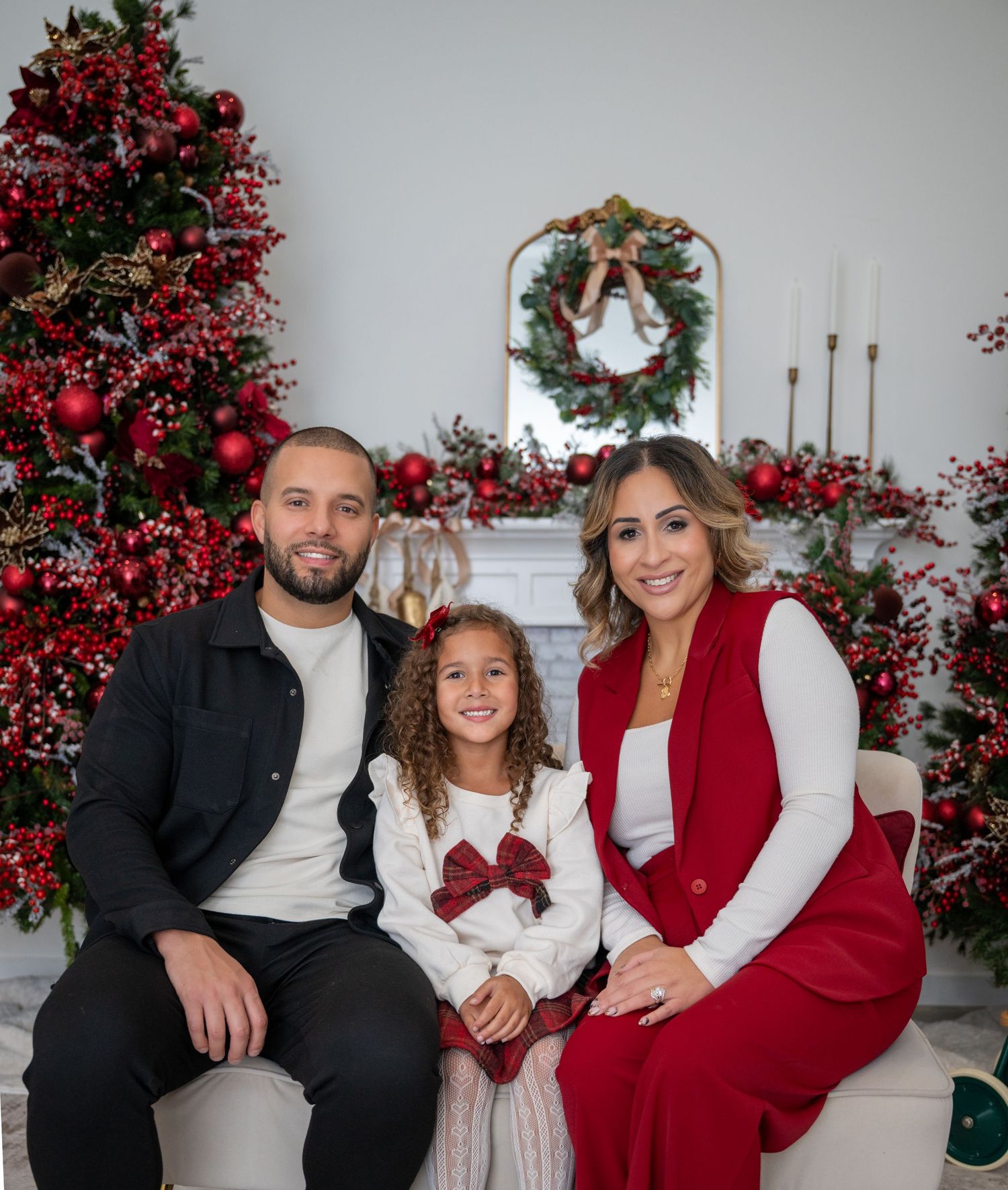 Family of three sitting together on a couch in front of a red Christmas tree and fireplace.