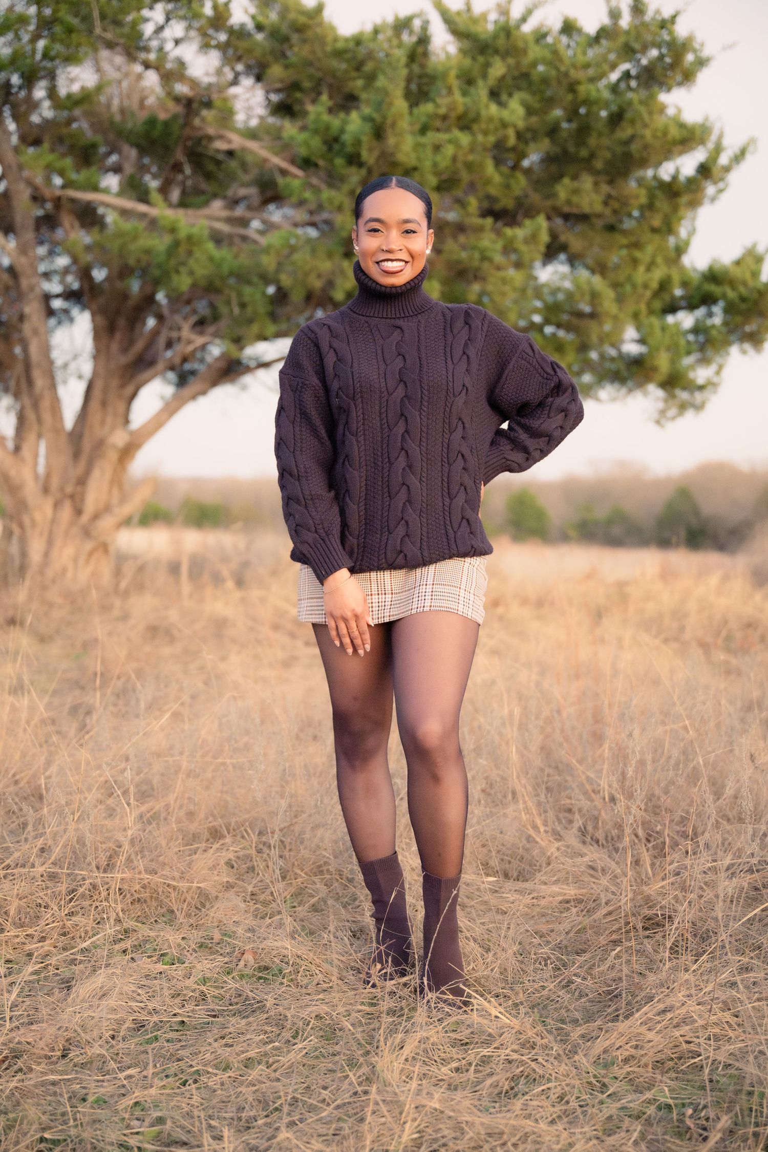 Smiling young Black woman in navy sweater and plaid skirt posing in an open field.
