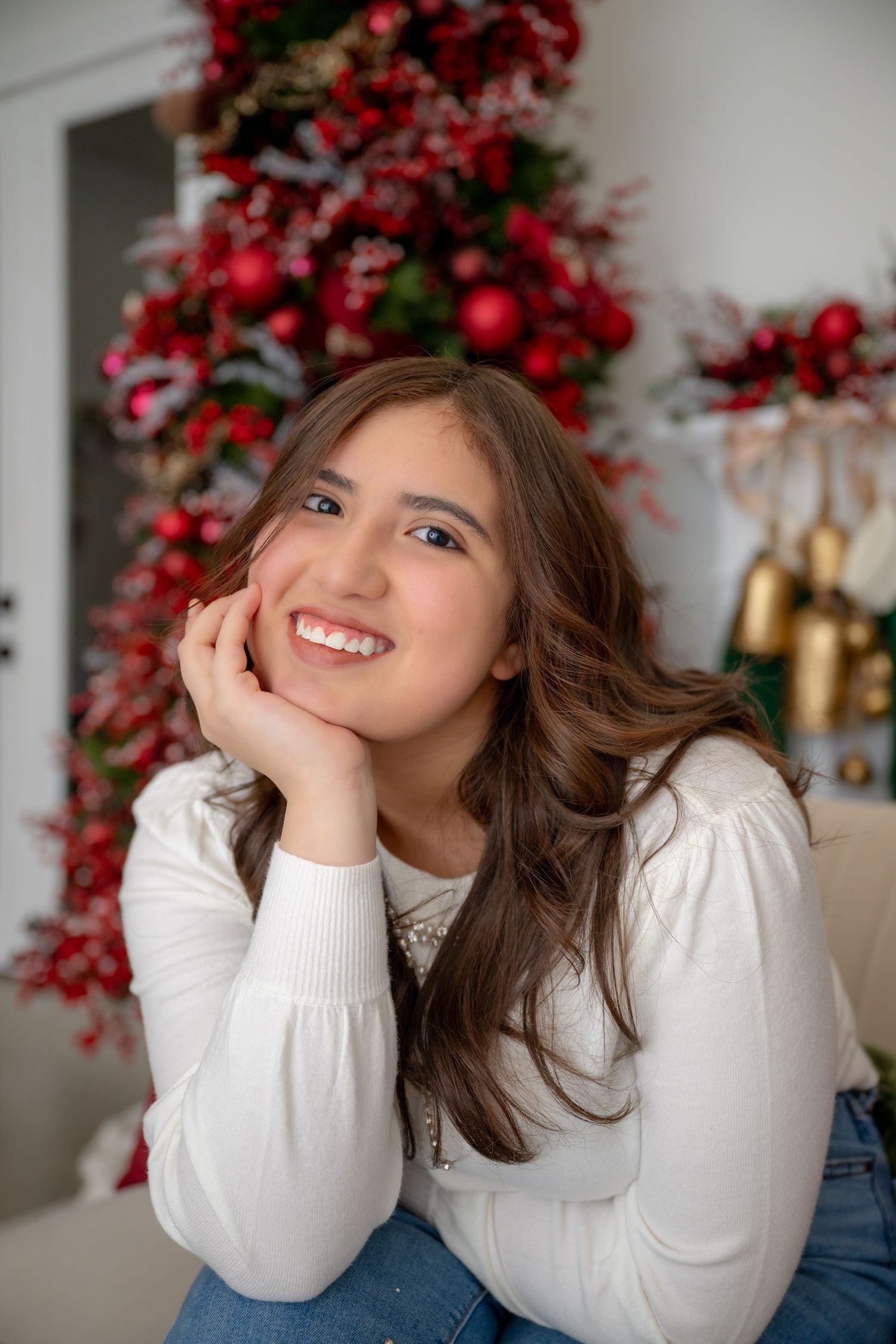 Smiling Senior in white sweater poses in front of a decorated Christmas tree with red ornaments.