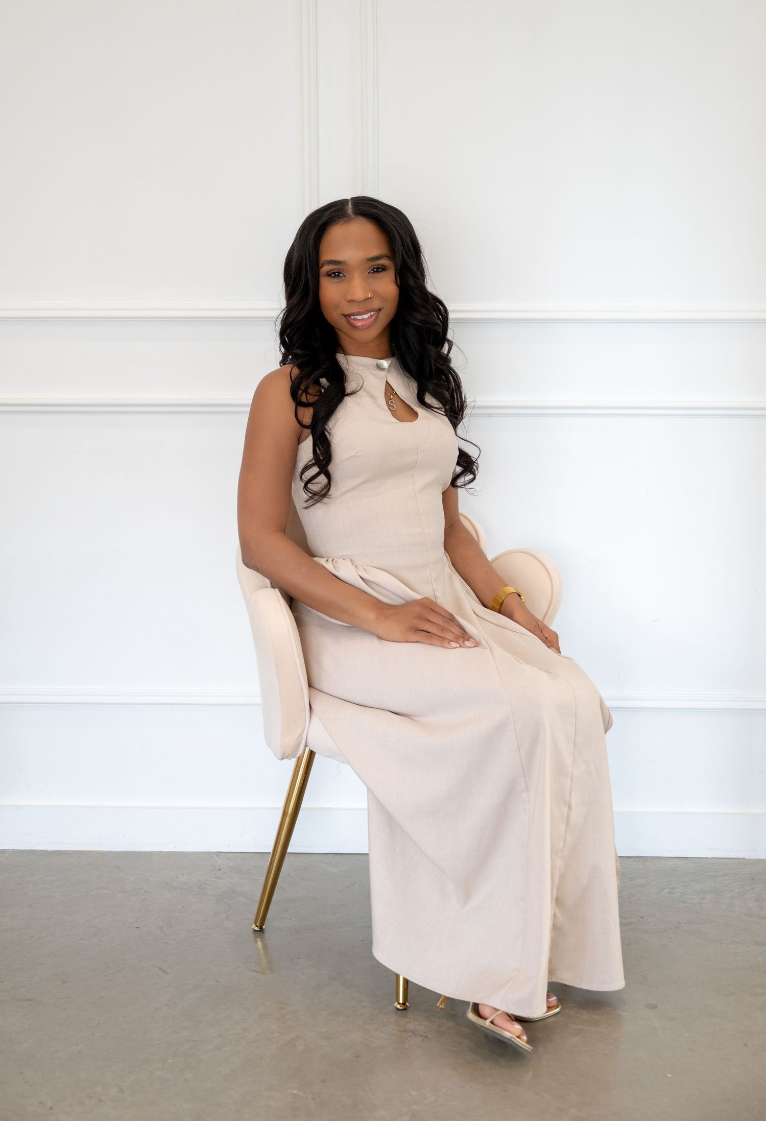 A woman in a beige sleeveless jumpsuit sits elegantly on a velvet chair in a bright white studio.