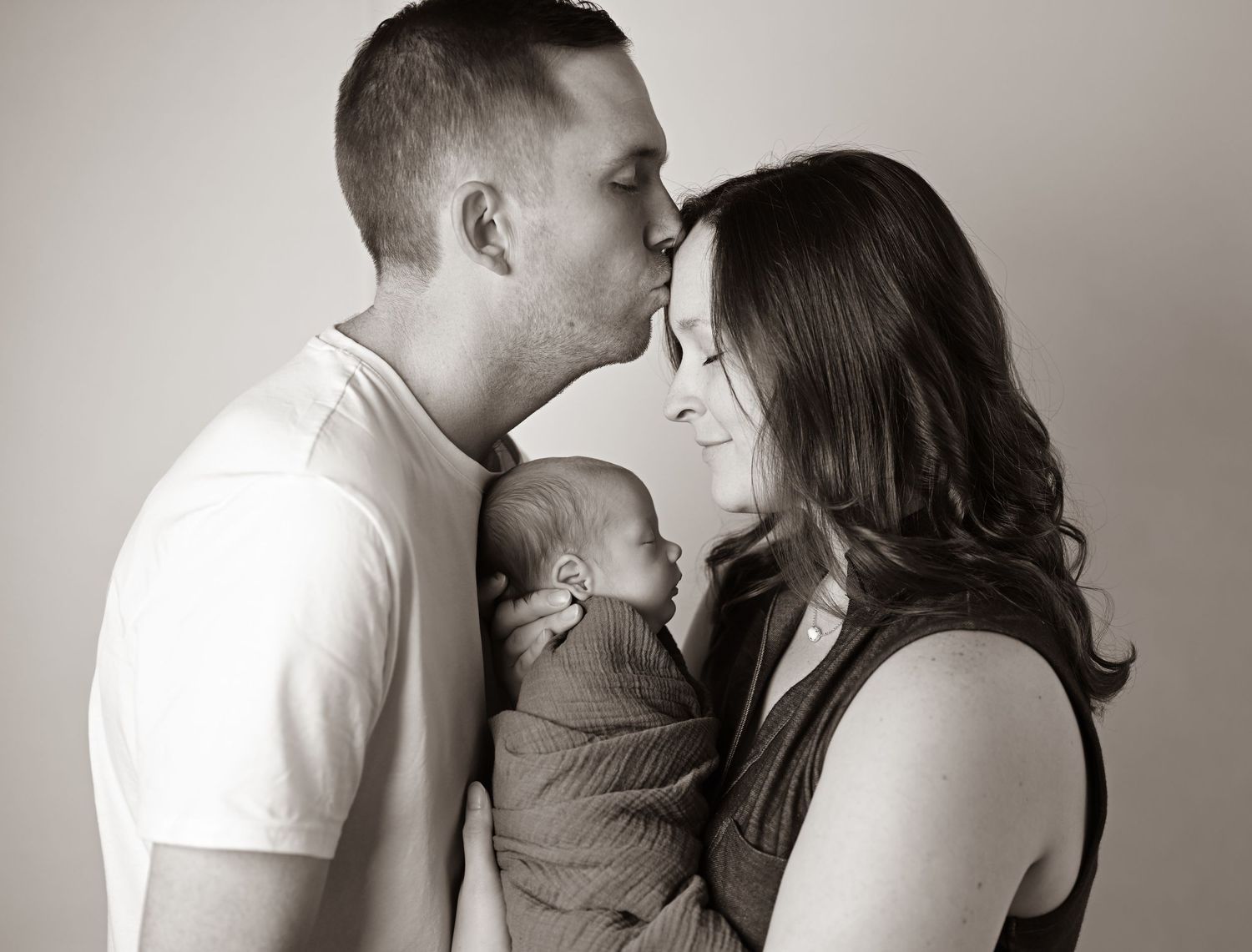 Black and white photo of a couple sharing a kiss over their newborn baby wrapped in a gray swaddle.