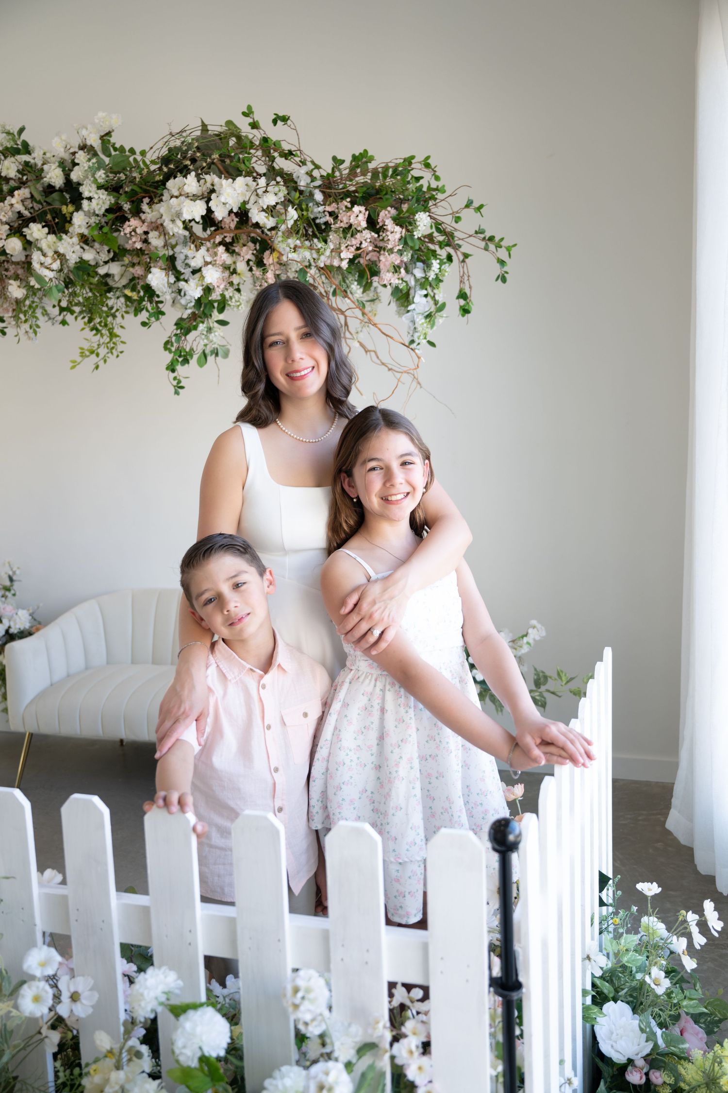 A mother and two children smile together before a white floral arrangement in a bright studio setting.