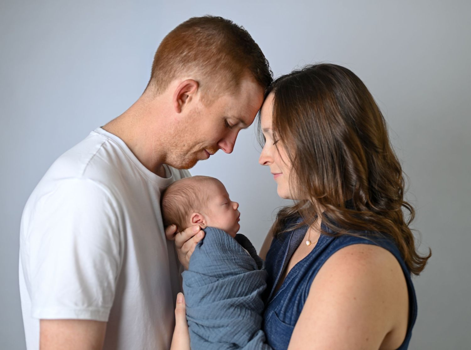 New parents tenderly hold their swaddled newborn baby together, touching foreheads in an intimate studio portrait.