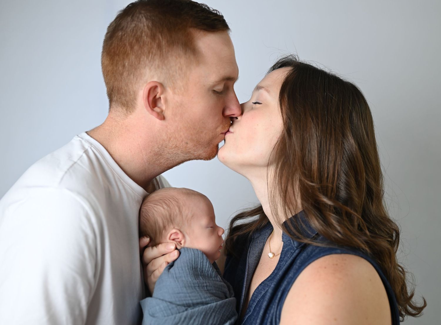 A couple shares a tender kiss while holding their newborn baby wrapped in a blue blanket during a family portrait.