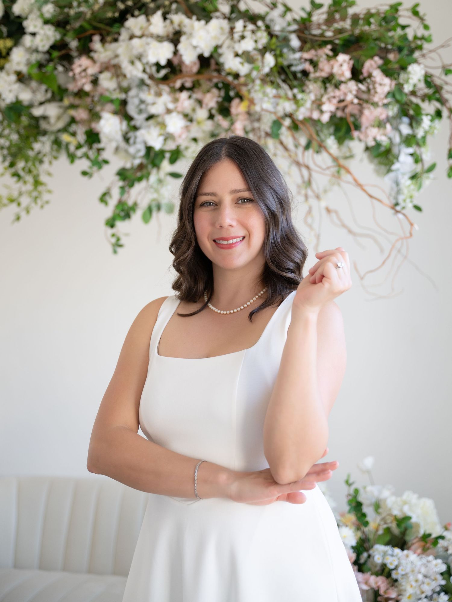 Smiling brunette woman in white square-neck dress with pearl necklace under pastel floral arrangement.