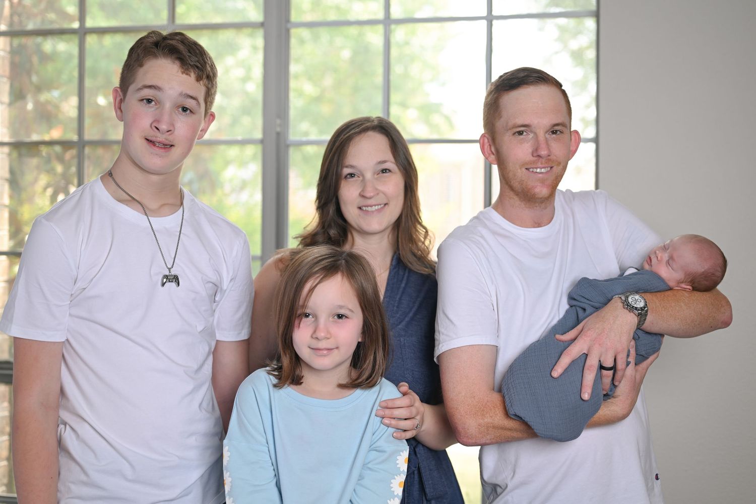 Family of five posing indoors, including two children, parents, and a newborn baby wrapped in a gray swaddle.