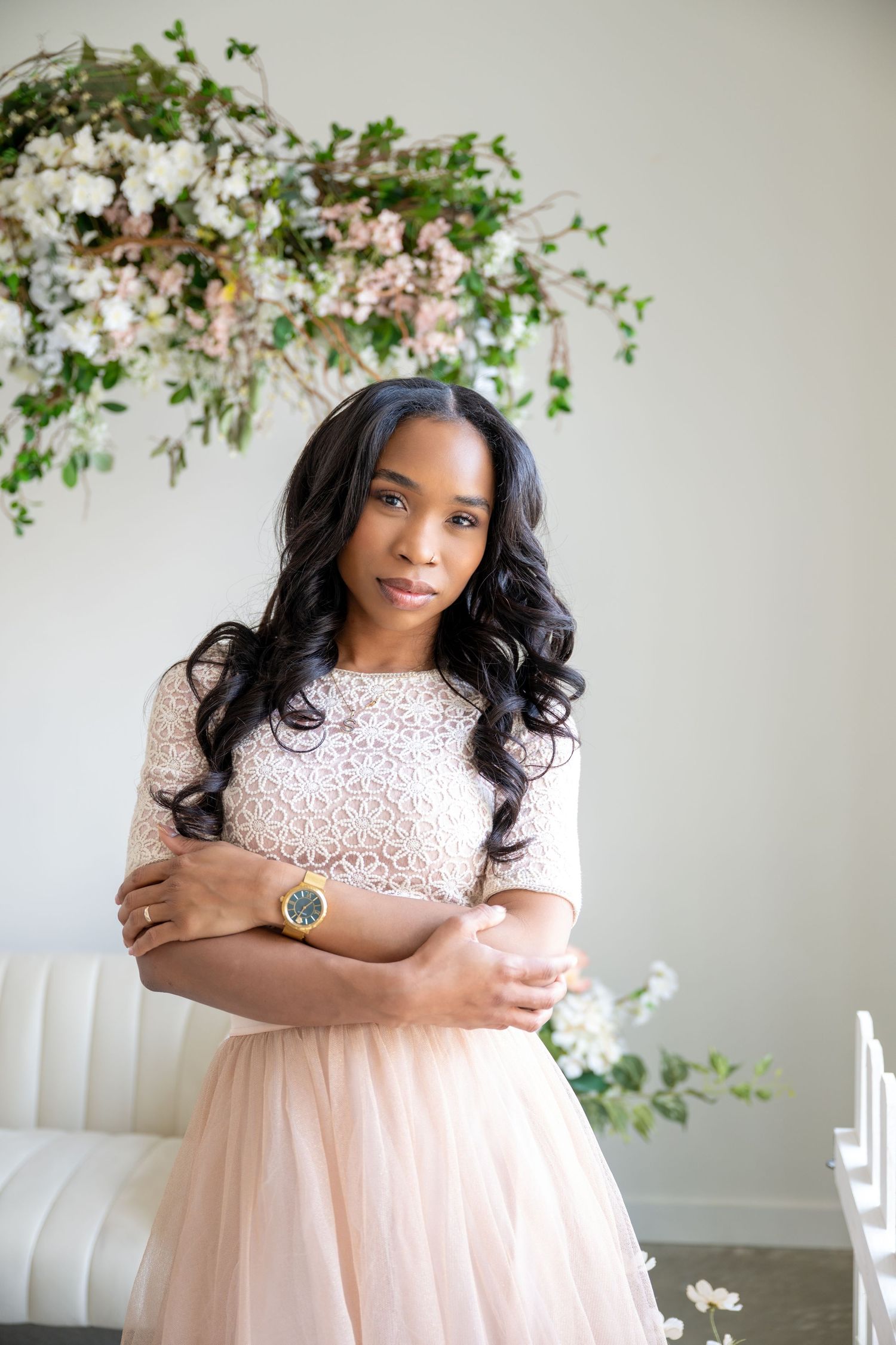 A woman in a white lace crop top and blush tulle skirt stands before a floral arrangement indoors.