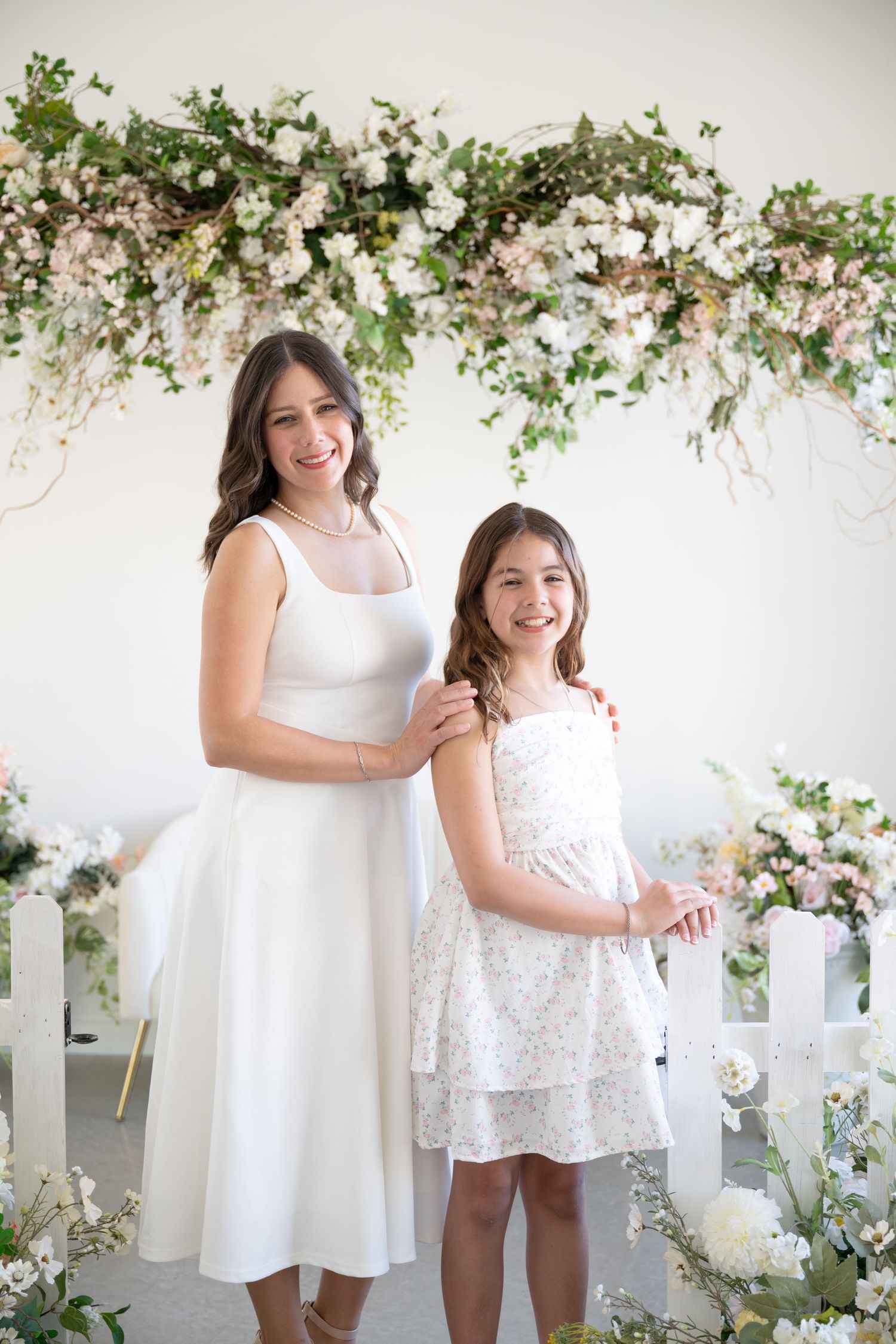 Woman and girl in white dresses posing under floral arch with pink and white blooms.