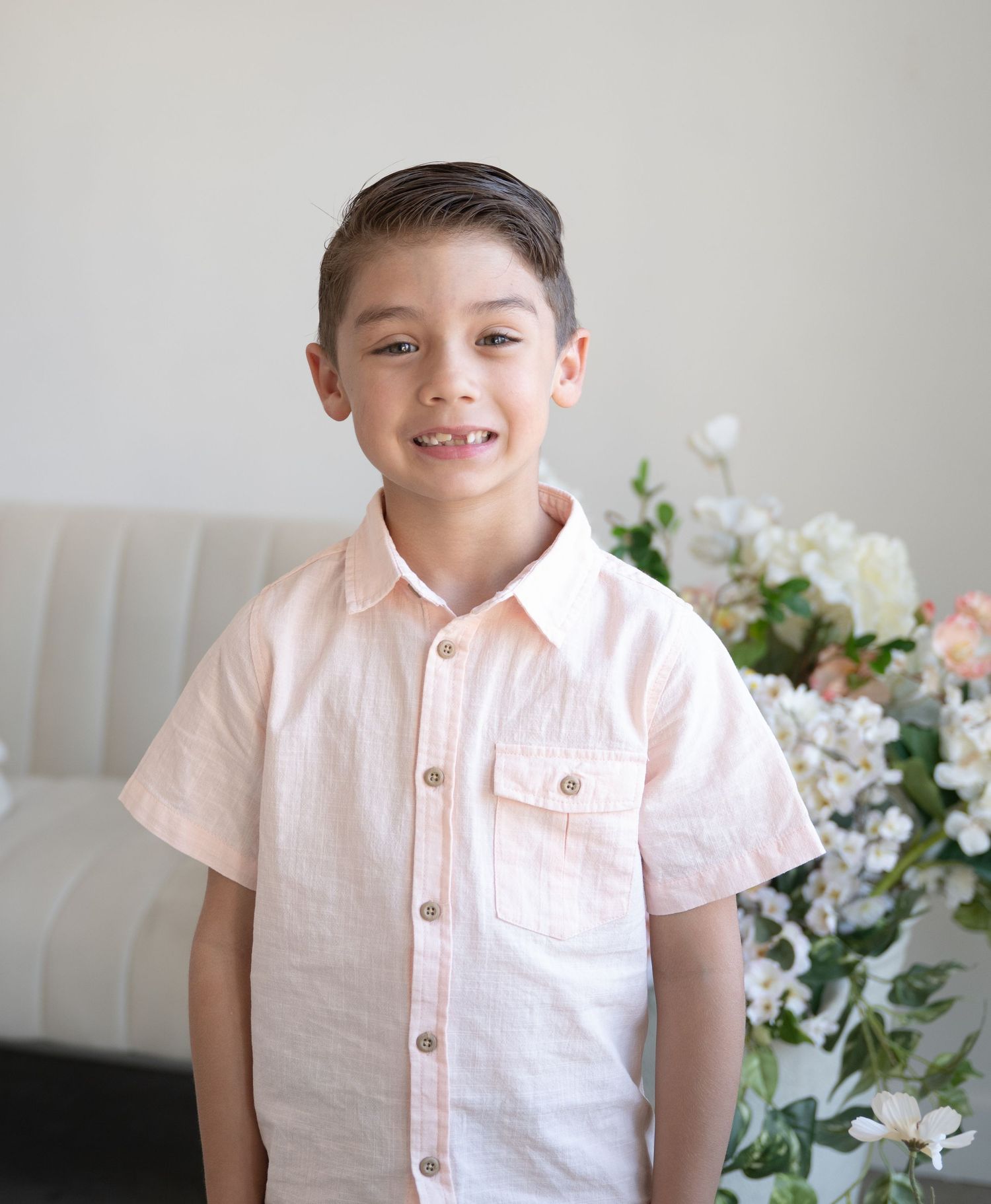 Smiling young boy in peach button-up shirt posing before floral arrangement in bright studio.