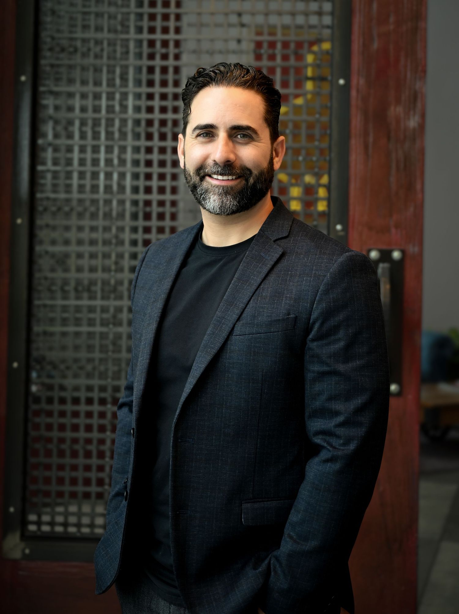 Smiling man with beard wearing navy blazer poses in front of decorative metal screen door.