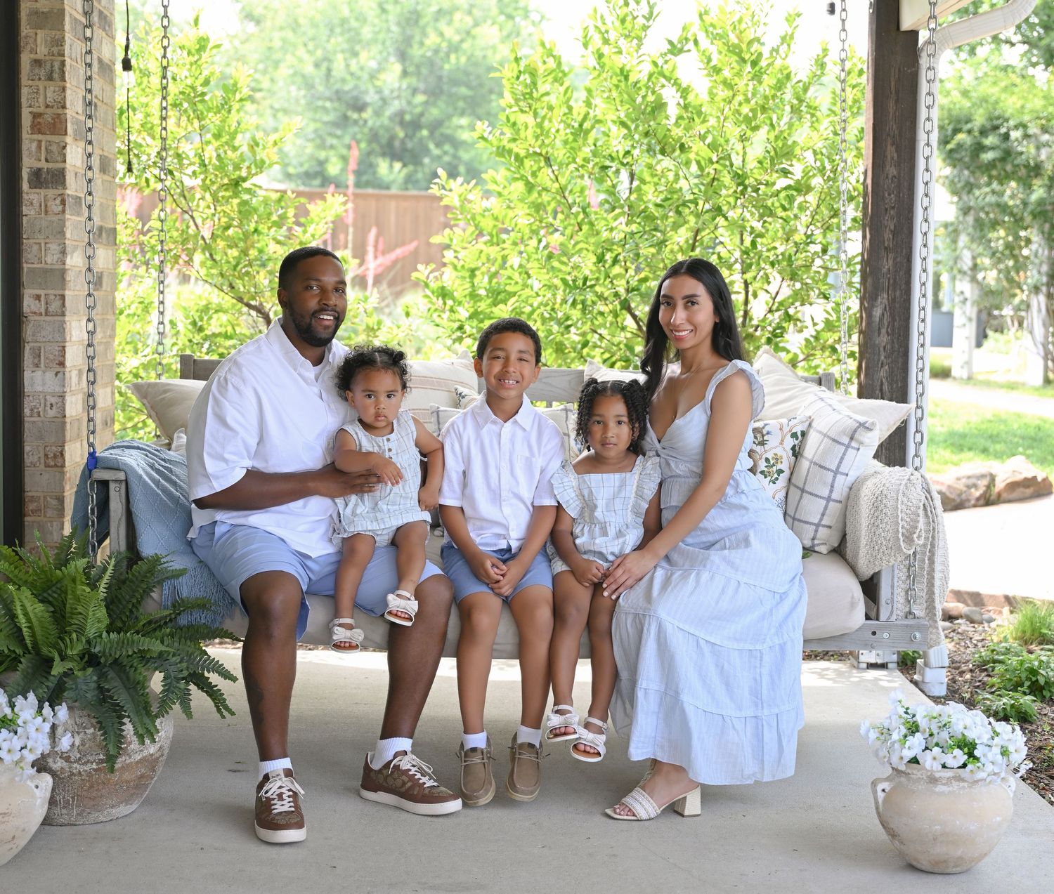 Family of five posing on a porch swing, dressed in white outfits, surrounded by lush greenery.