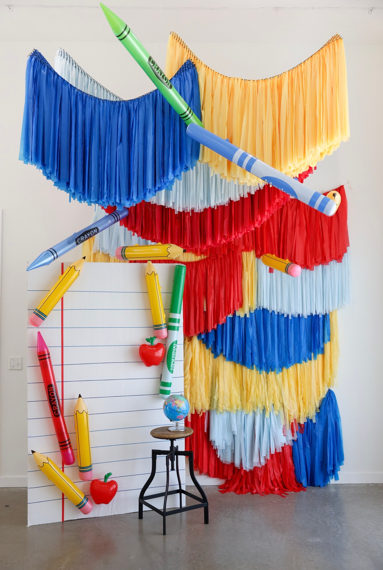 Colorful back-to-school photo backdrop with giant inflatable crayons, pencils, and fringe streamers in red, blue, and yellow.
