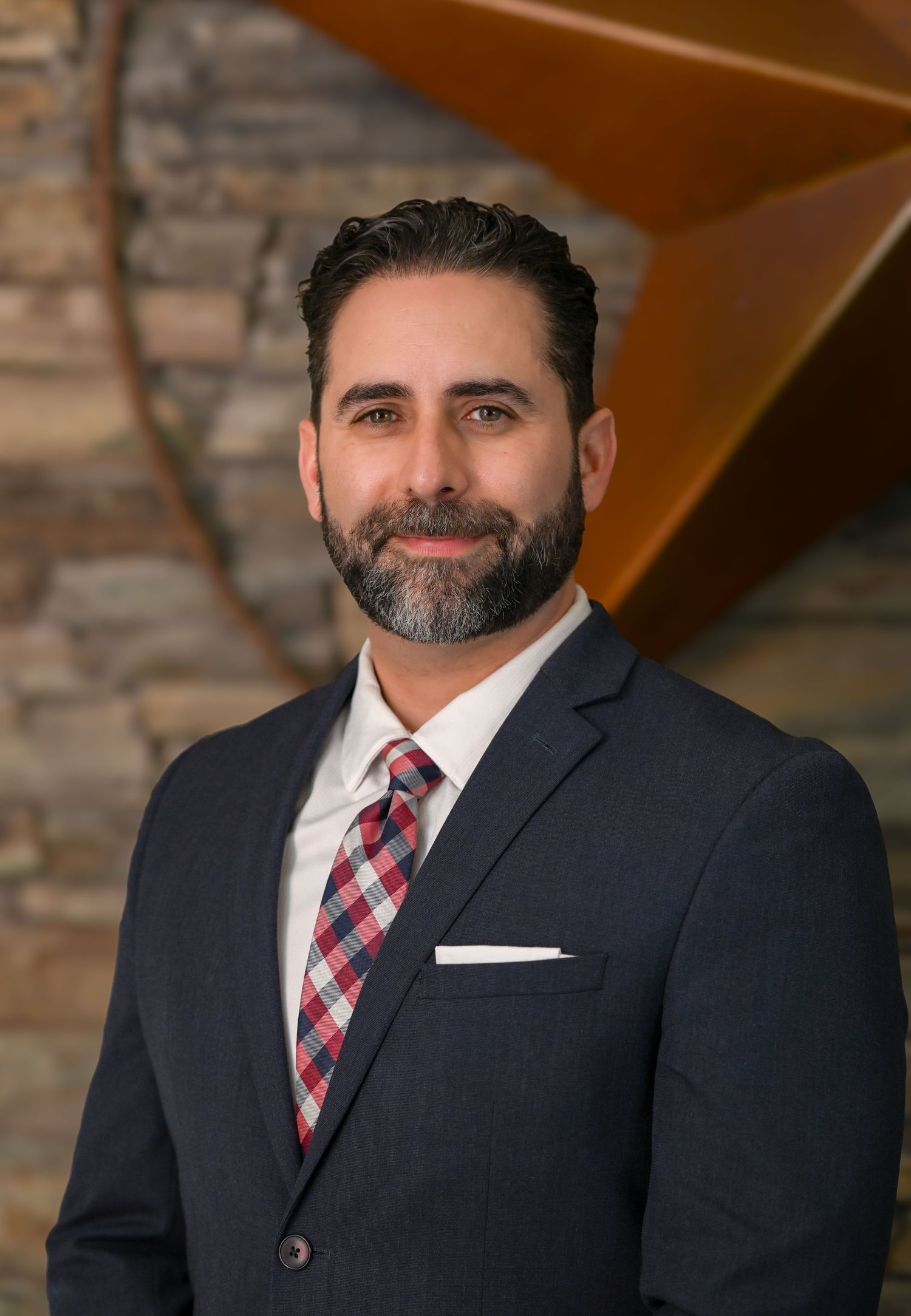 Professional man in navy suit and plaid tie poses for corporate headshot in modern office setting.