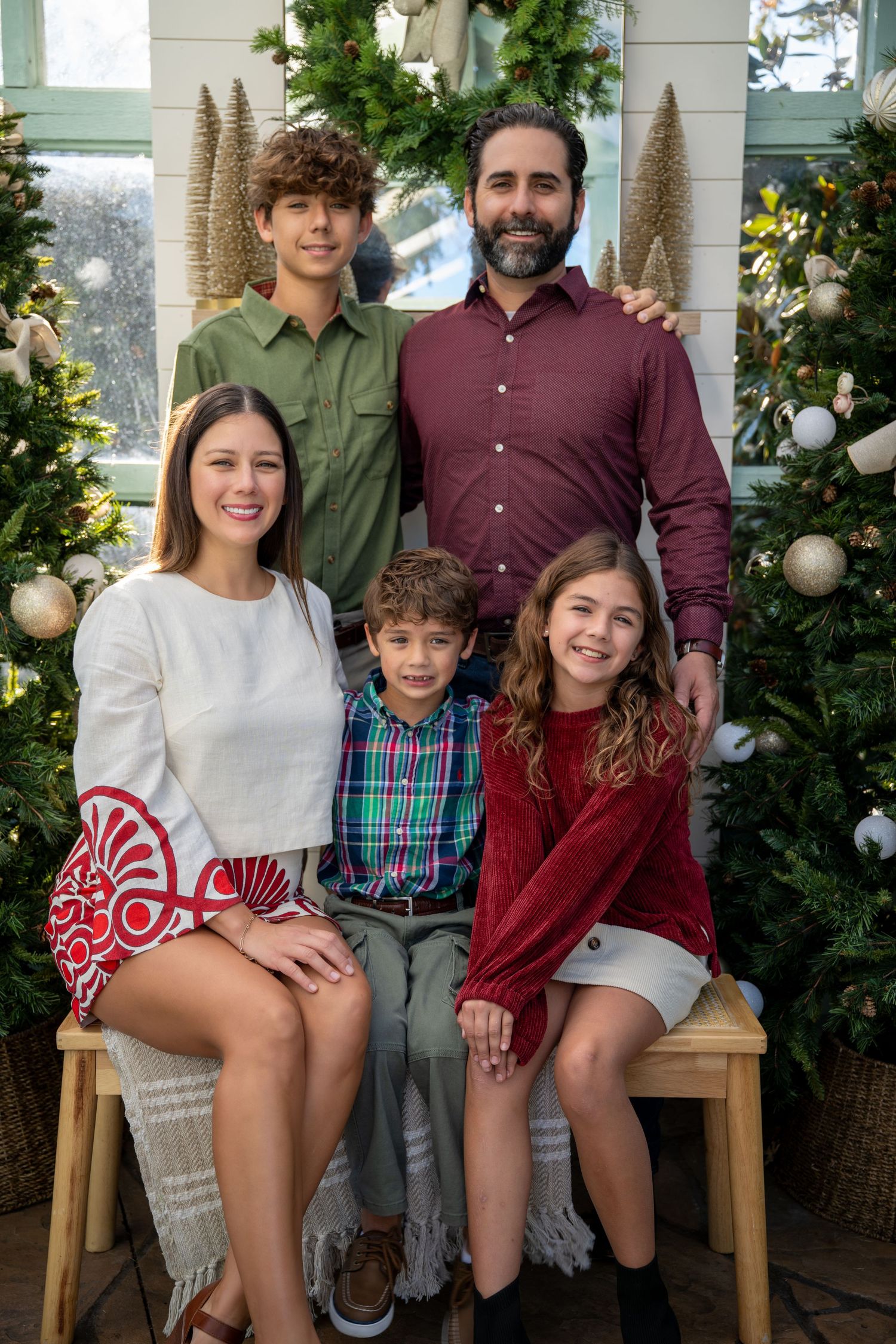 Family of five smiling together inside a festive greenhouse decorated with Christmas trees.