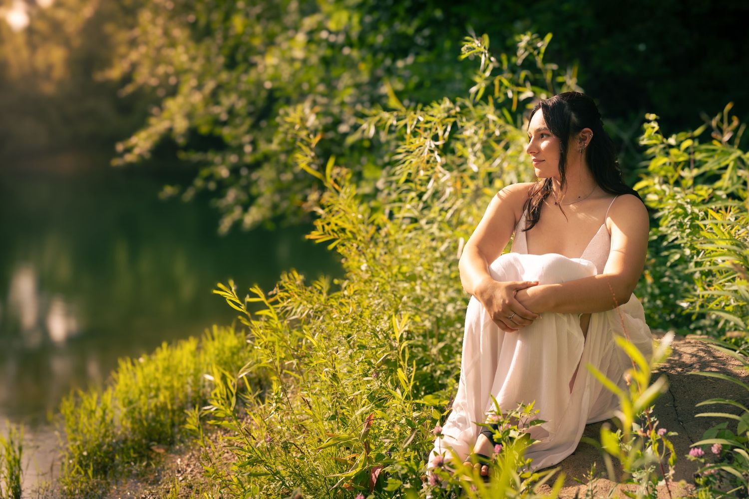 A woman in a white dress sits peacefully by a sunlit pond surrounded by lush green trees and plants.