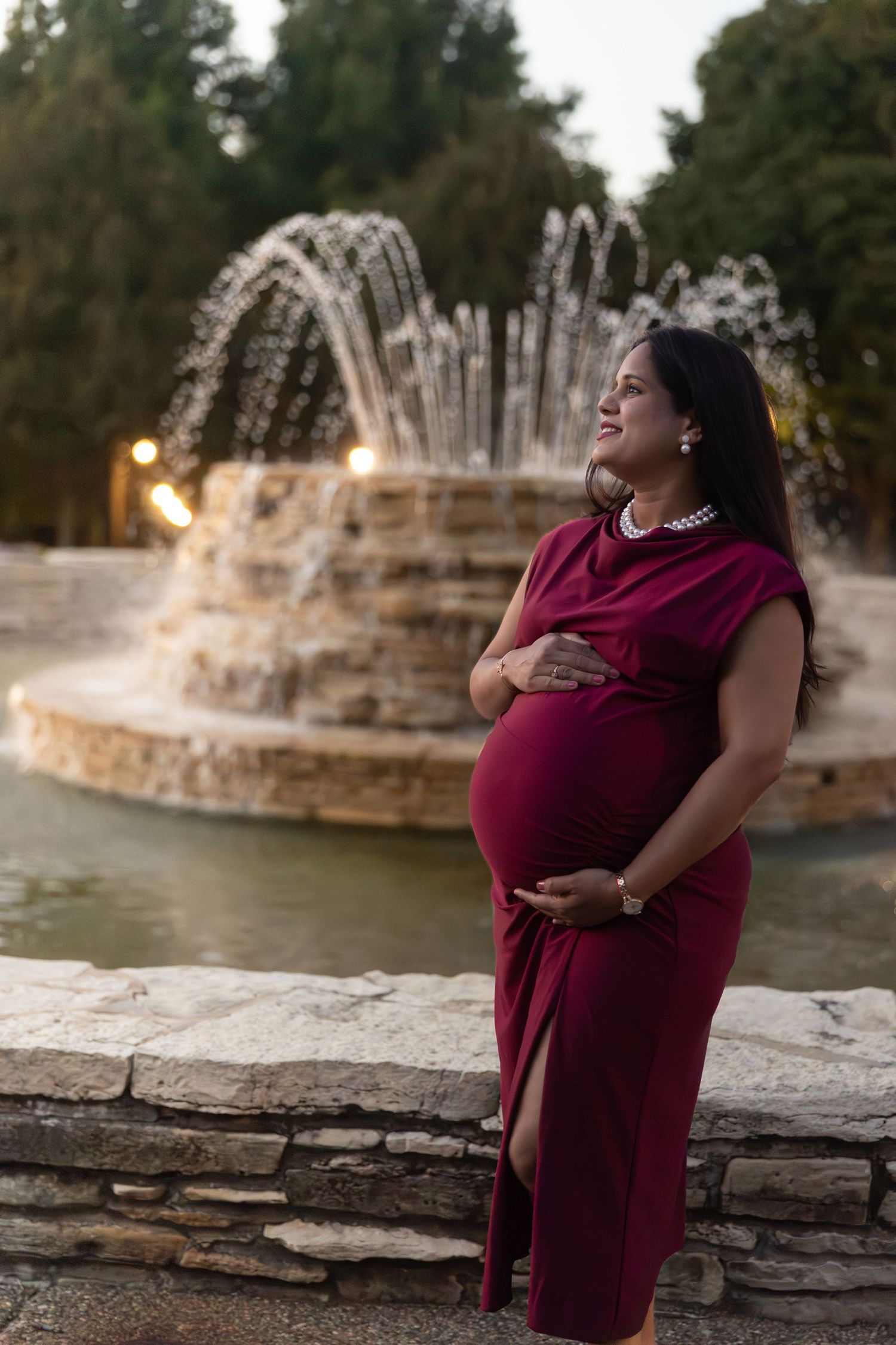 Pregnant woman in burgundy dress cradles baby bump by stone fountain at dusk.