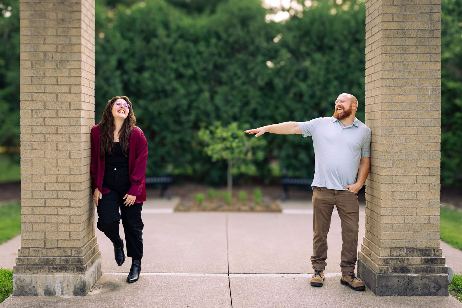 A man and woman laugh between brick pillars outdoors, man pointing humorously while woman leans back smiling.