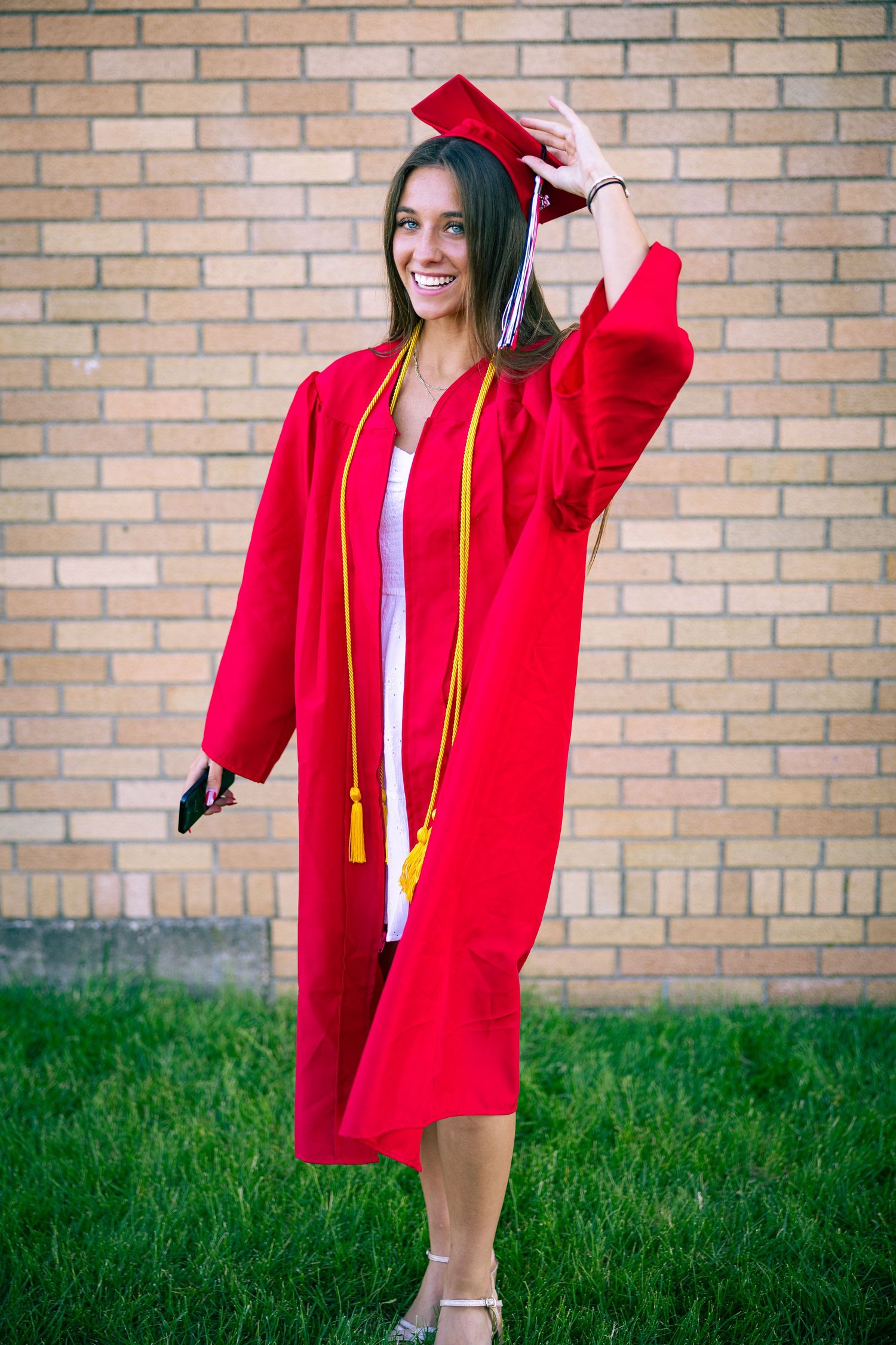 Graduate in red cap and gown poses against brick wall outdoors.