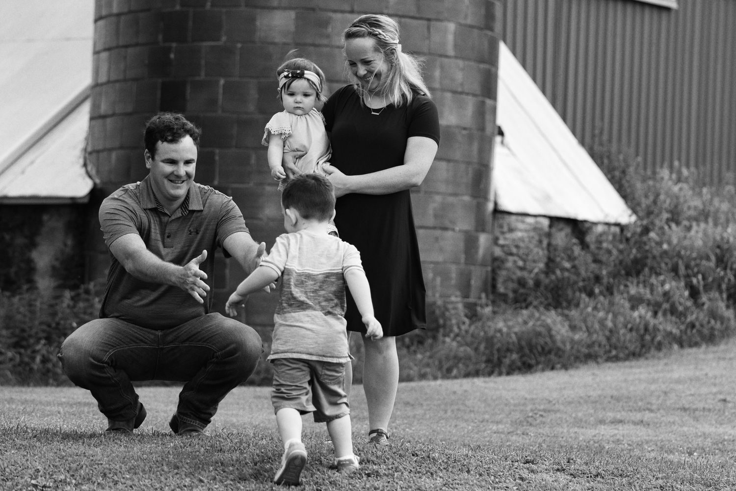 Black and white photo of a young family outdoors, father crouching as toddler walks toward him, mother holding baby near a barn.
