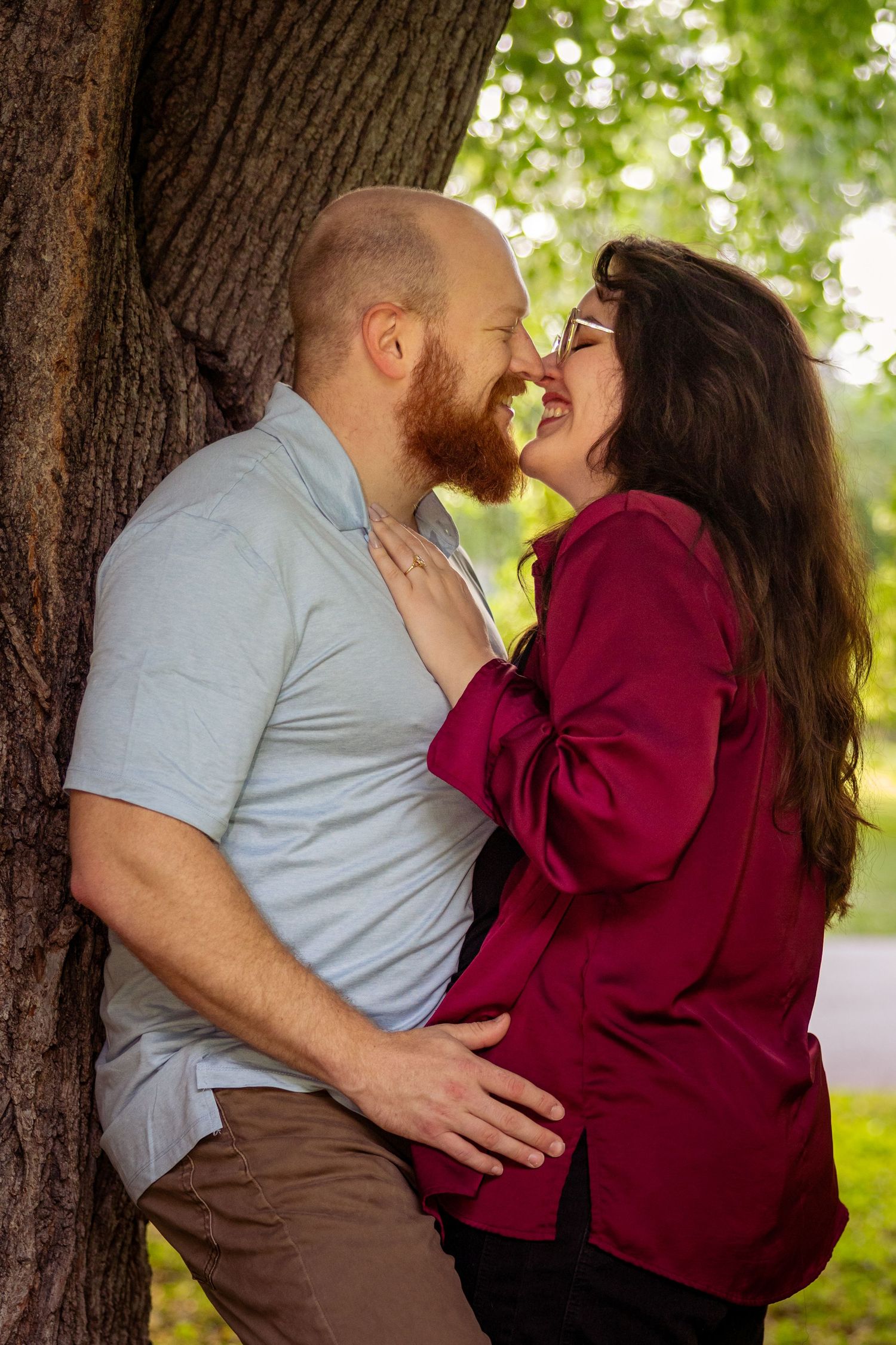 A couple sharing a romantic moment leaning against a tree in a lush green park, smiling and touching noses.