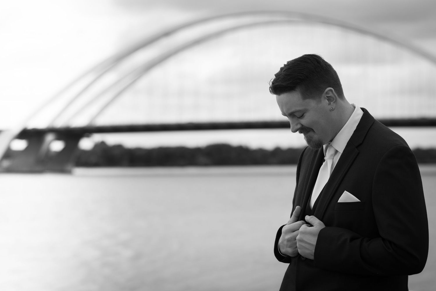 Black and white photo of a man in a suit adjusting his jacket near a river with an arch bridge in the background.