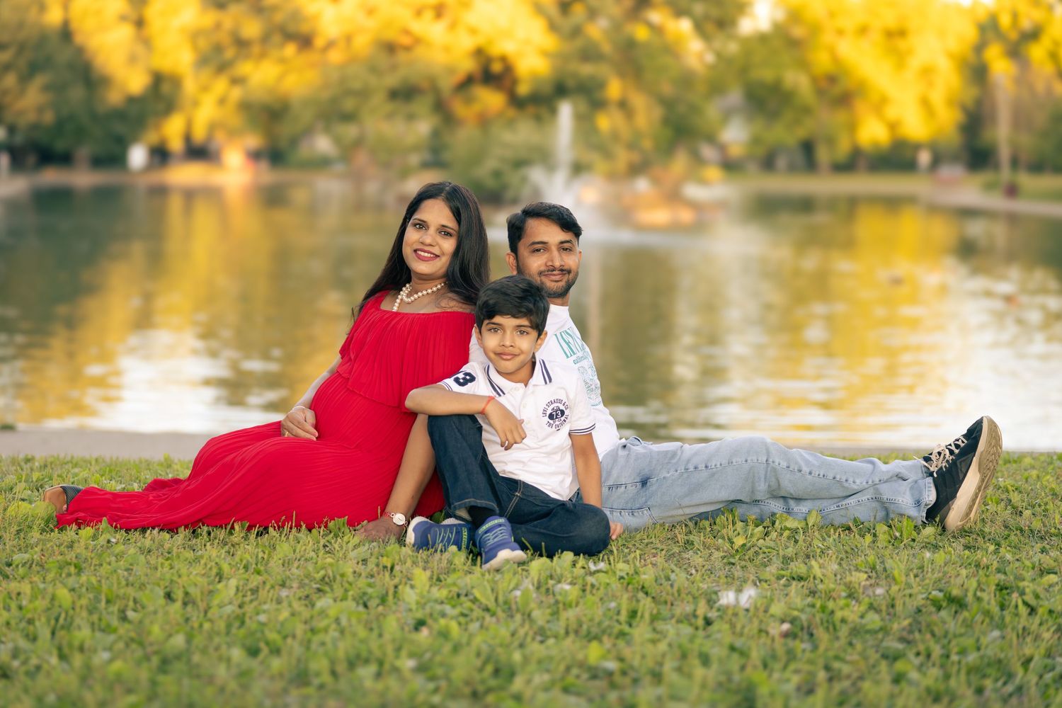 A family sits together on grass near a pond with vibrant yellow fall foliage in the background.