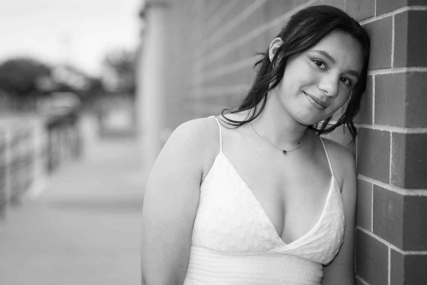Black and white portrait of a young woman in a white dress leaning against a brick wall, smiling softly.