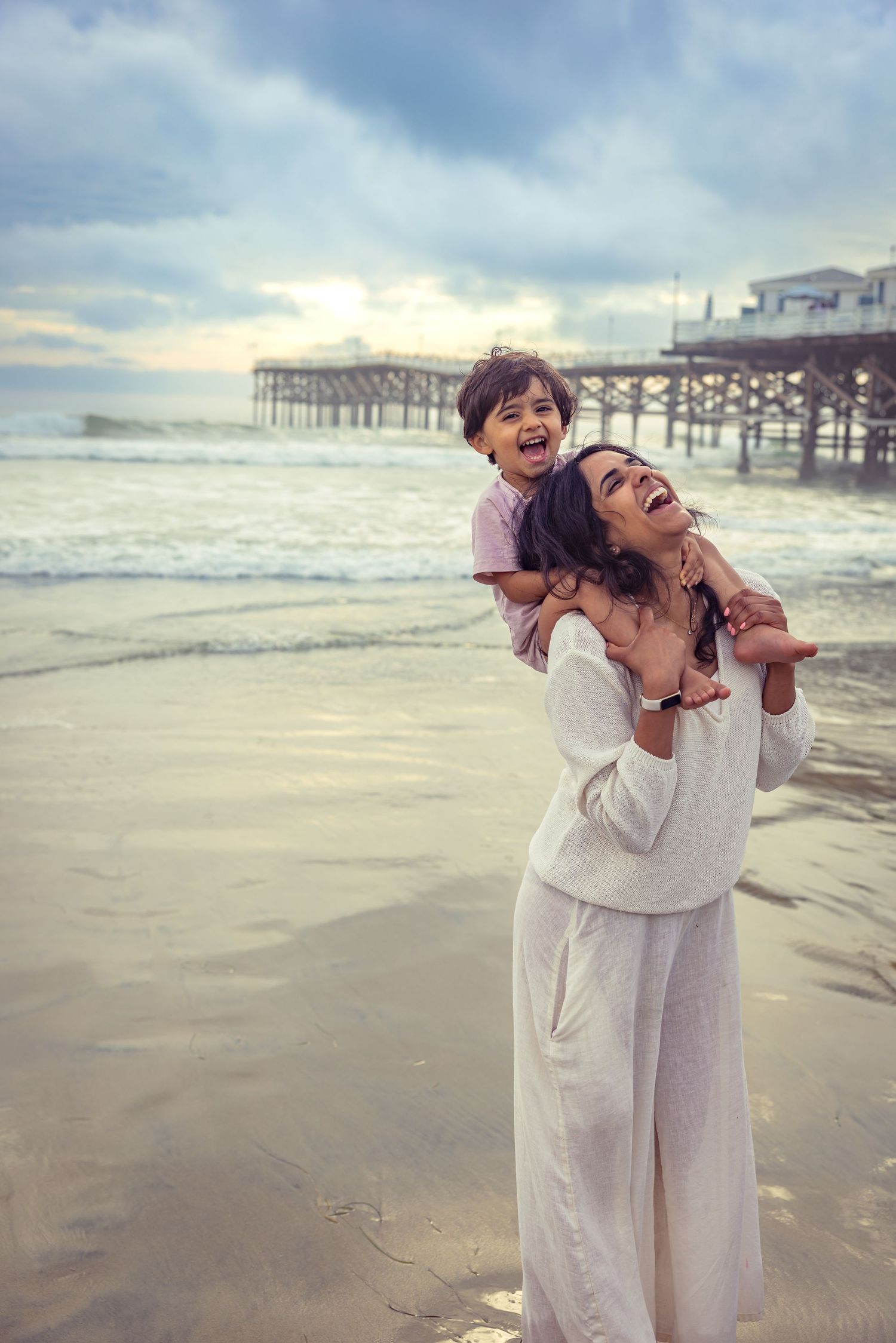 Couple embracing on a beach at sunset with a pier in the background.